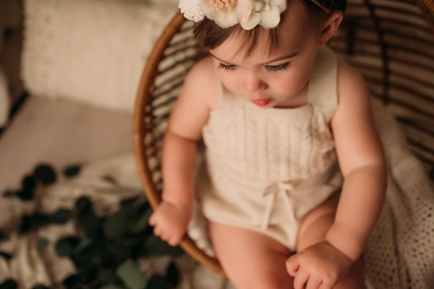 Baby in cream knit outfit looks down while sitting in rattan chair with floral accents.