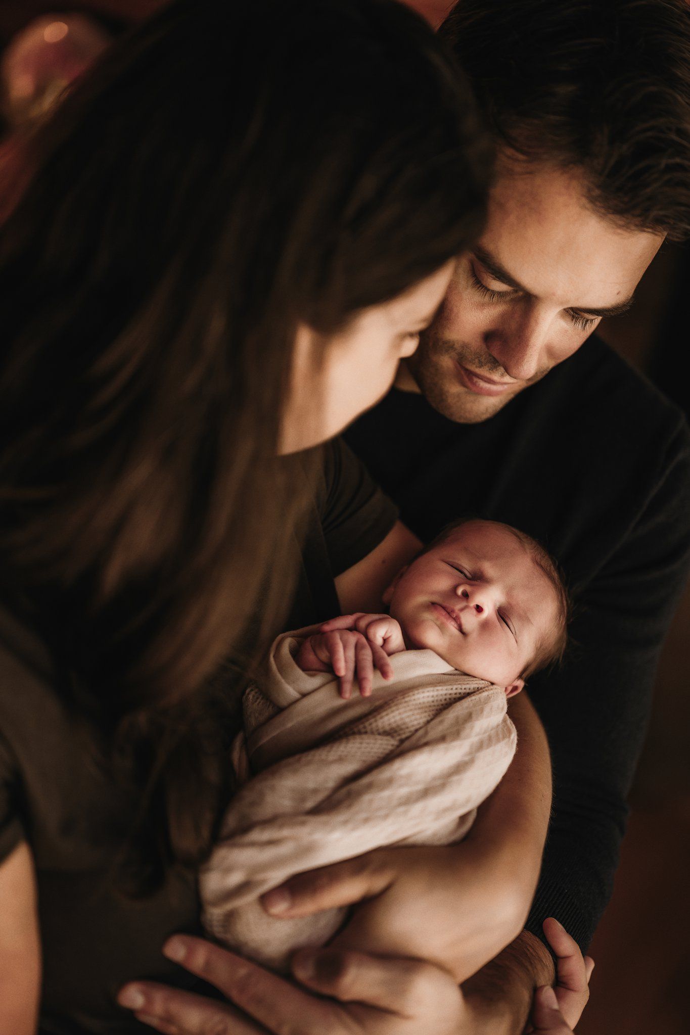 Intimate indoor family moment with newborn swaddled in beige blanket.