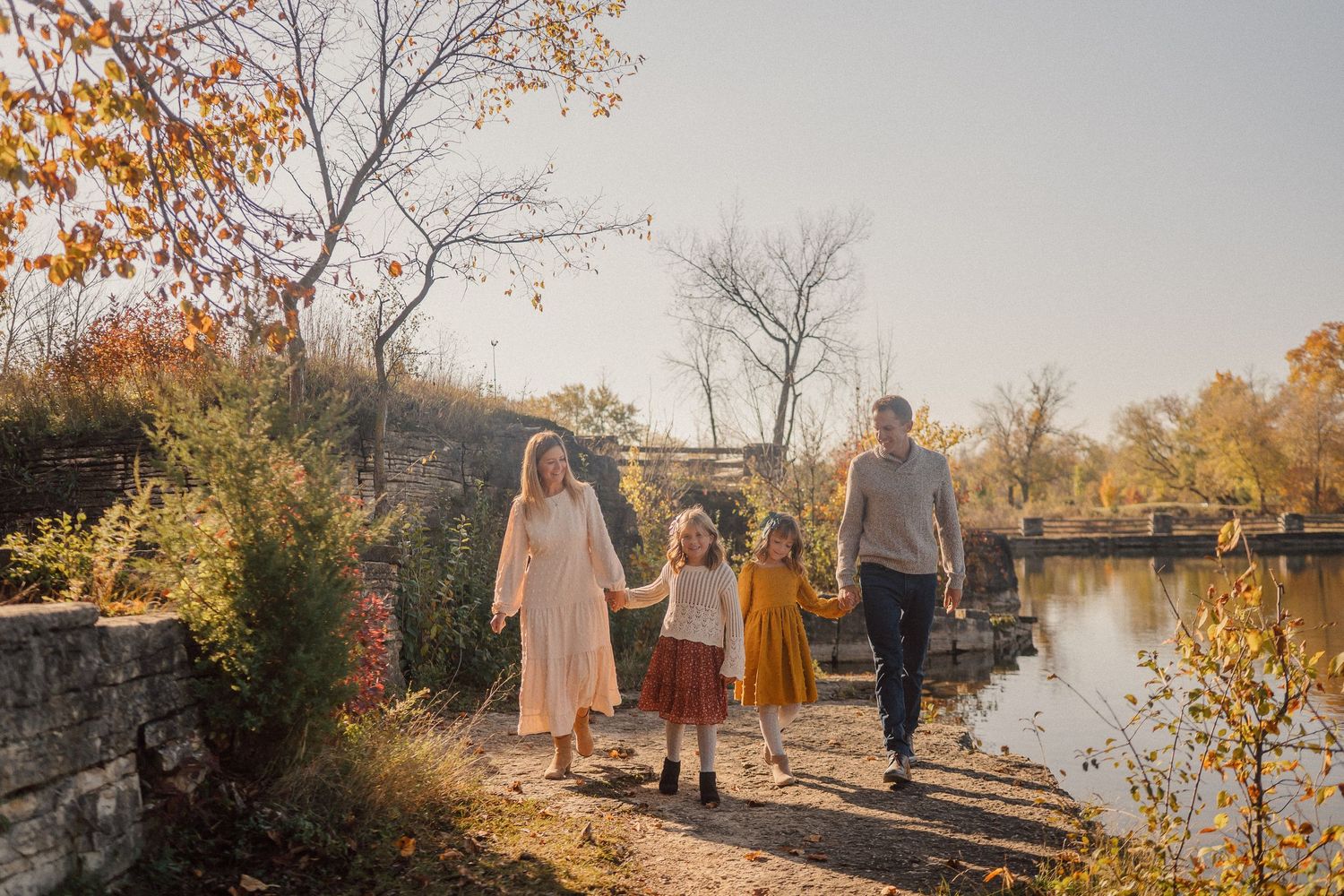 A family walks along a scenic stone path by a river during autumn with colorful fall foliage in the background.