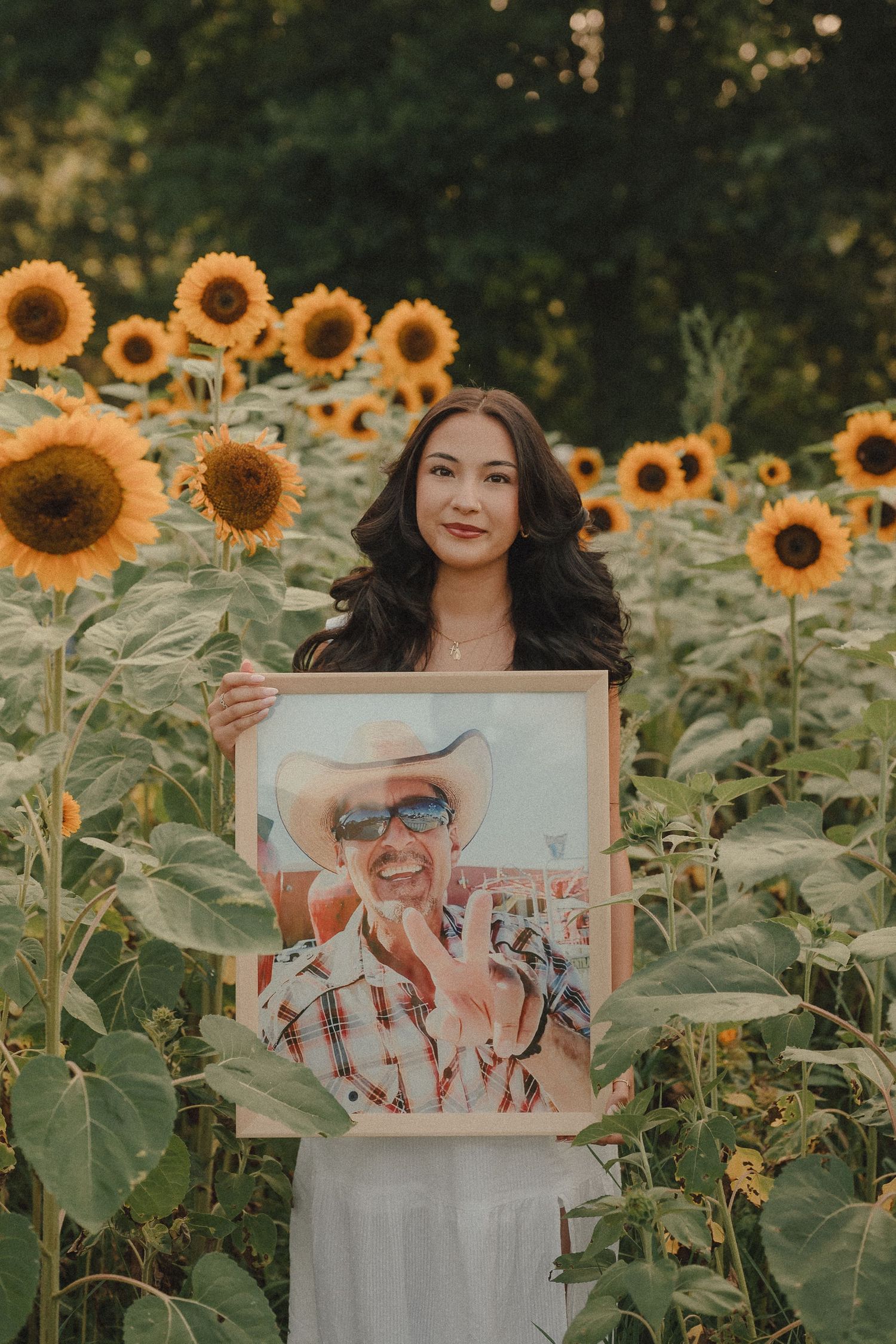 Young woman in white dress holding a framed portrait in a sunflower field.