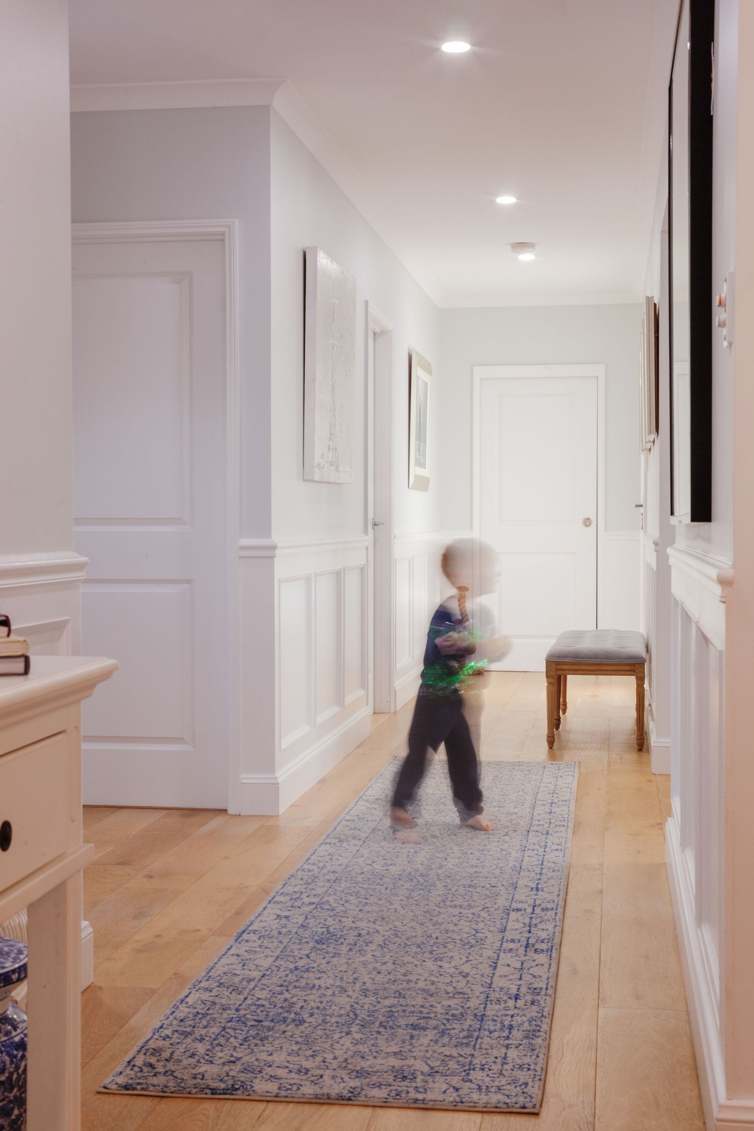 Sunshine Coast lifestyle photography: child running through a modern white hallway with wainscoting, timber floors, and a blue runner rug.