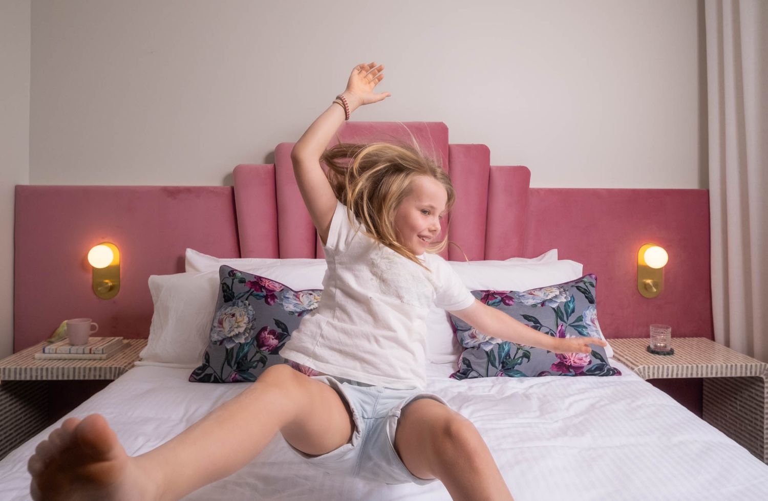Sunshine Coast lifestyle photography: young girl jumping on a luxury bed with a pink velvet headboard and gold sconce lighting