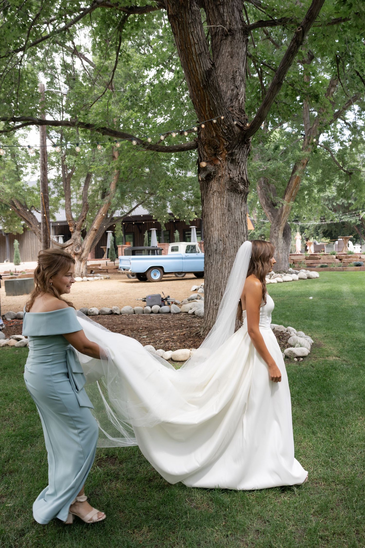 Bridesmaids in sage green dresses help carry a wedding dress train across green lawn.