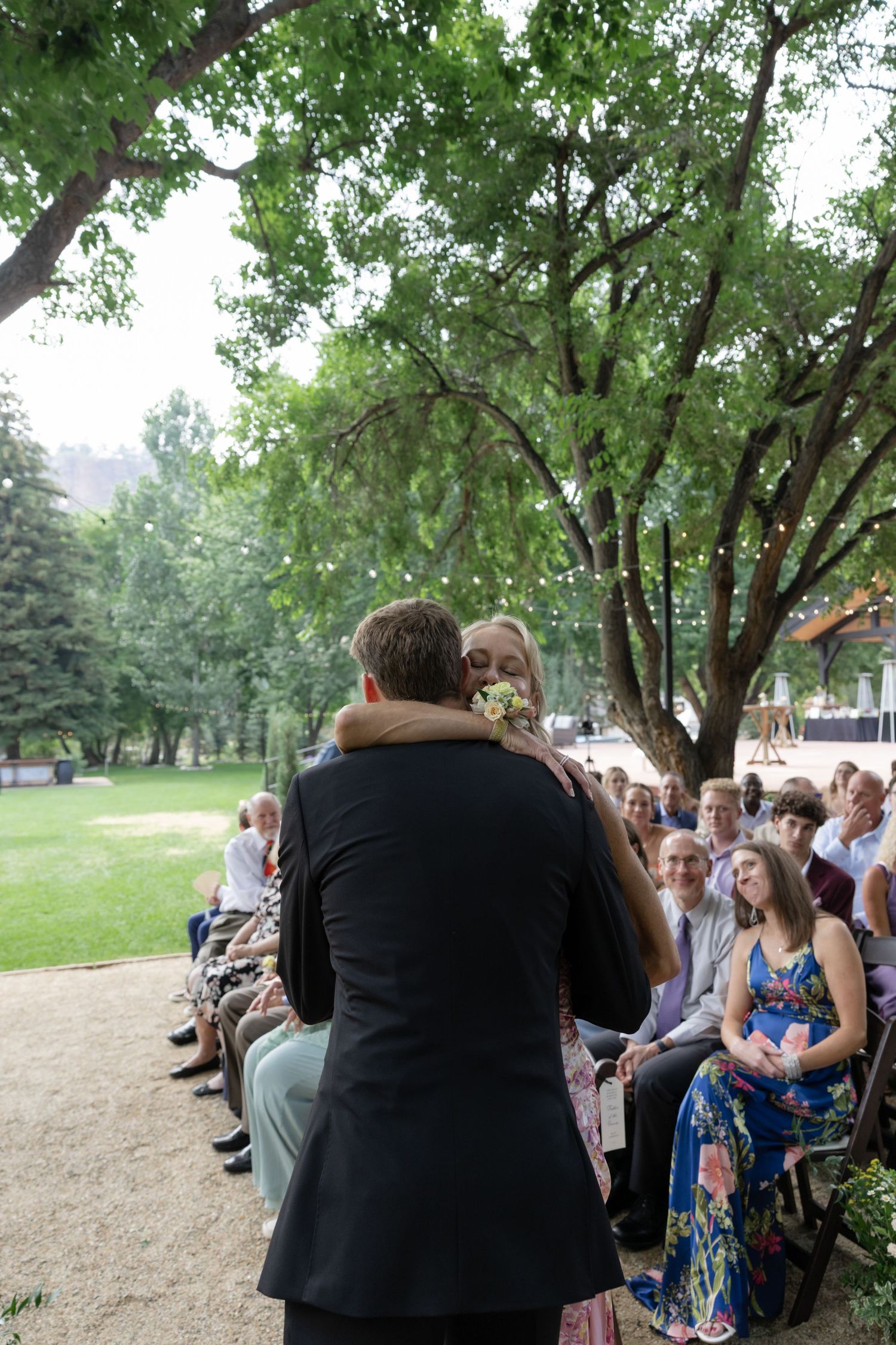 Wedding ceremony audience seated outdoors under large trees.