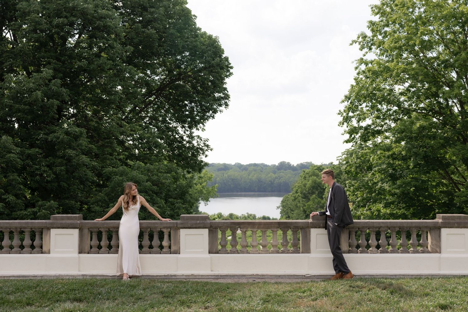 A couple stands apart on a stone terrace overlooking a serene lake view.