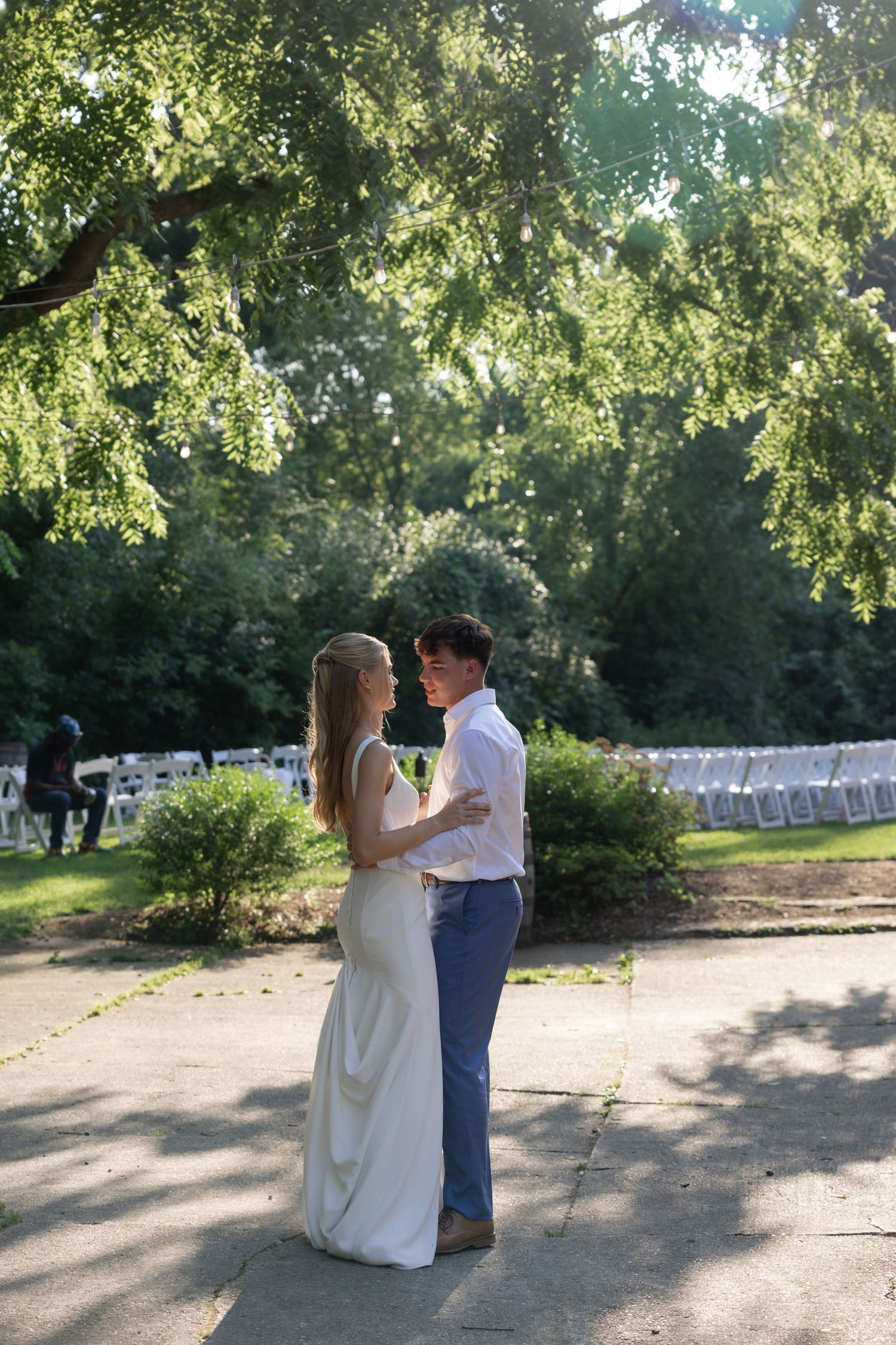 A couple shares a tender moment under sunlit trees at an outdoor wedding venue with rows of white chairs.