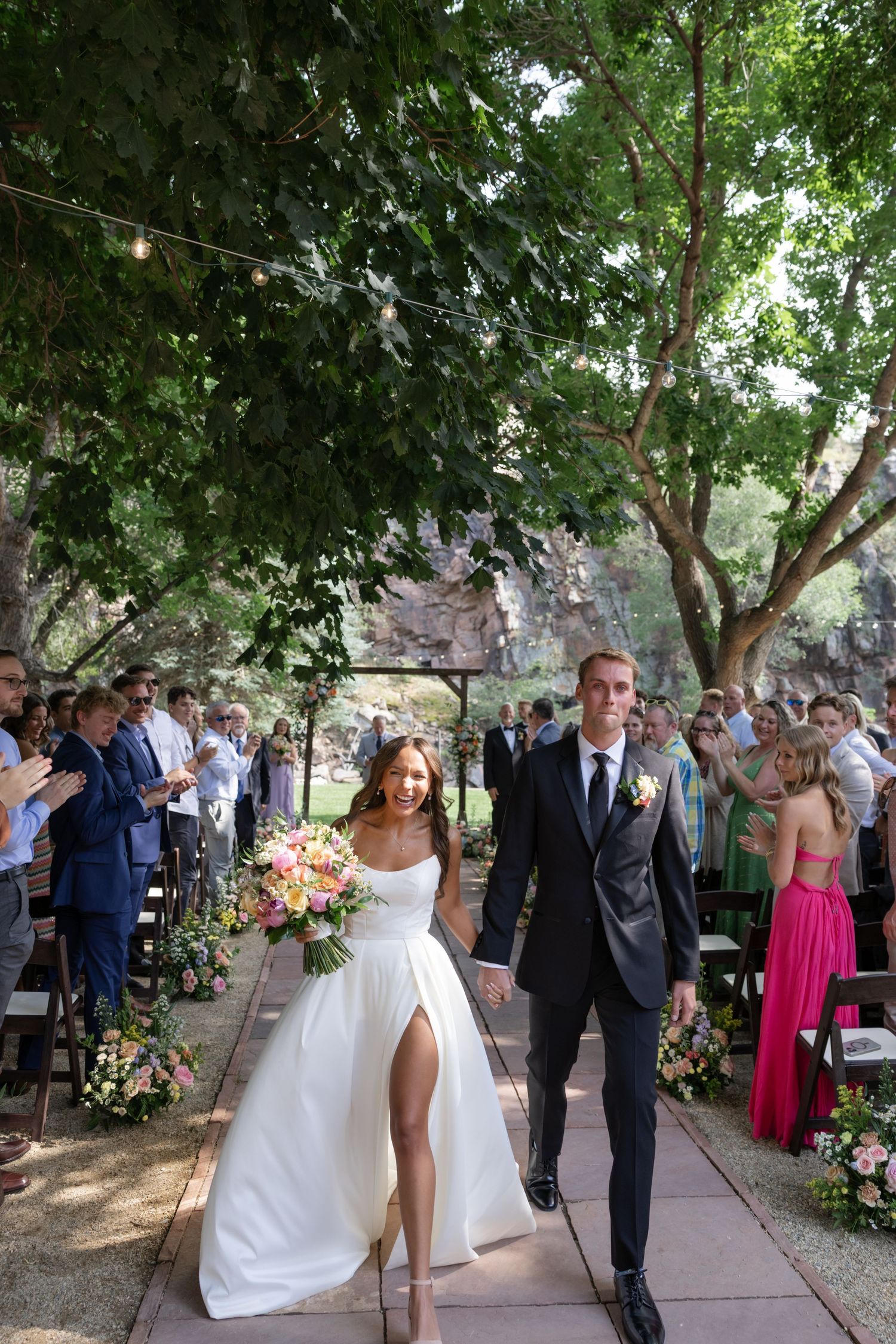 A couple walks down a flower-lined pathway after their outdoor wedding ceremony.
