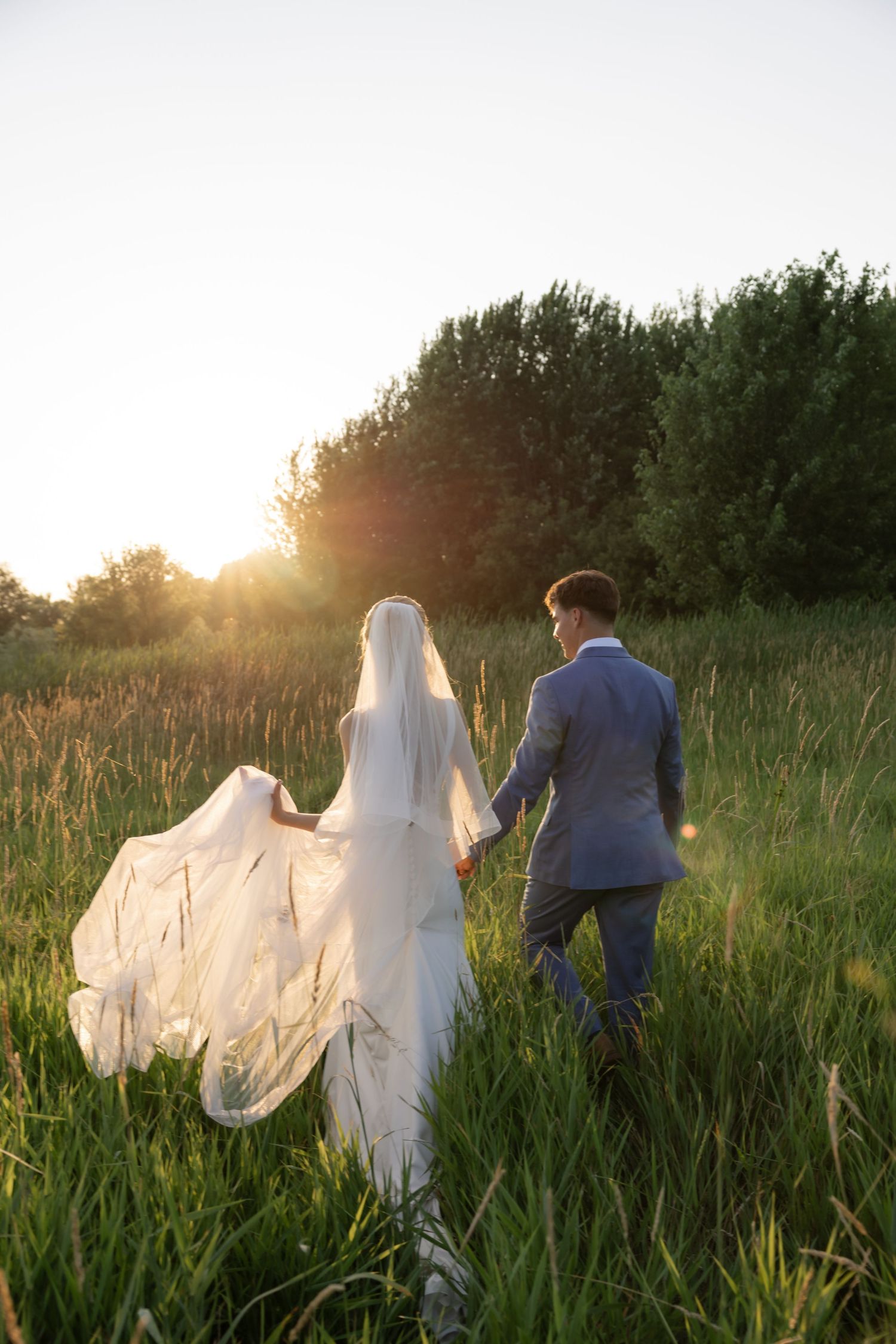 Bride and groom walking through tall grass at sunset with flowing veil.