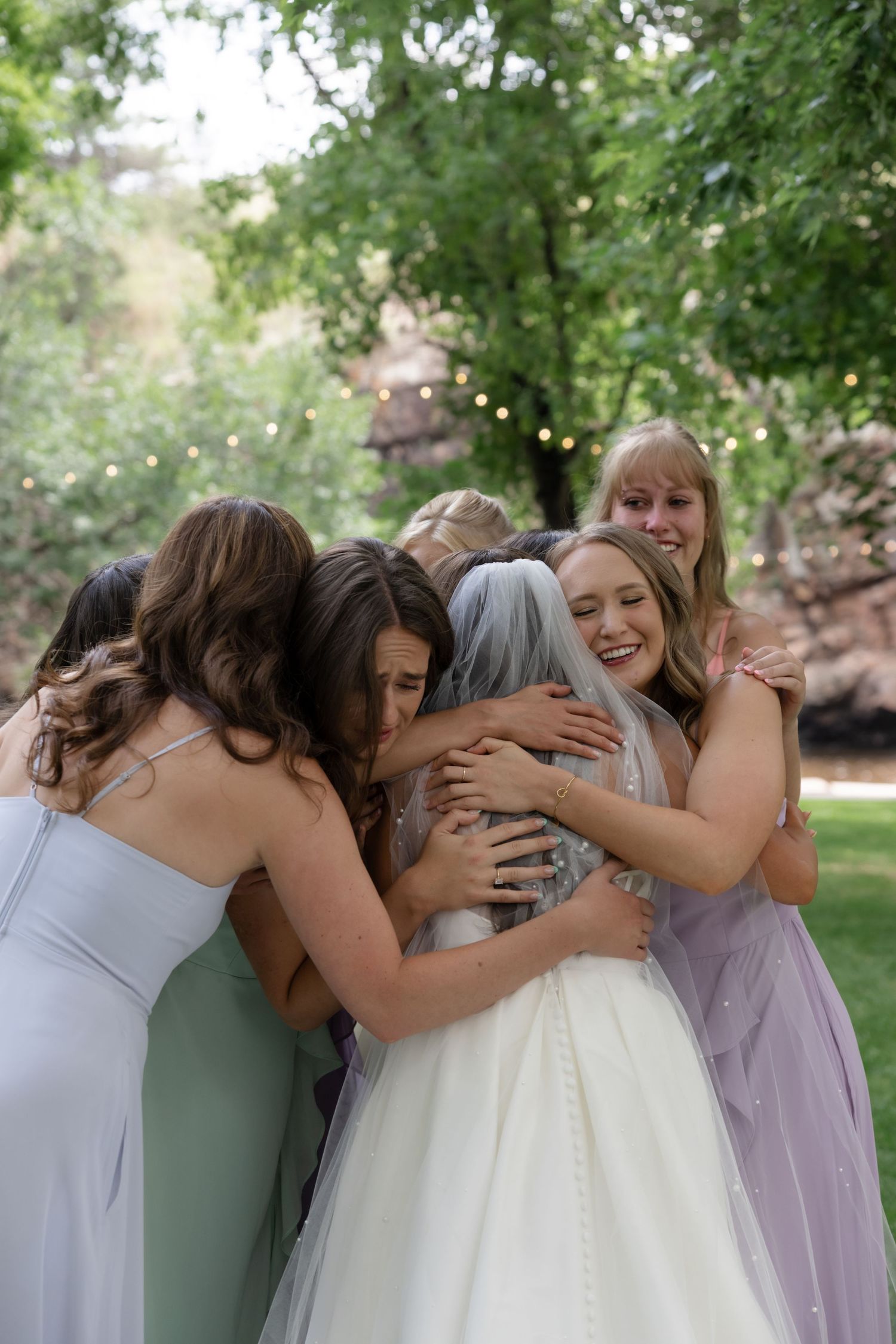 Bridesmaids in pastel dresses share an emotional group hug outdoors.
