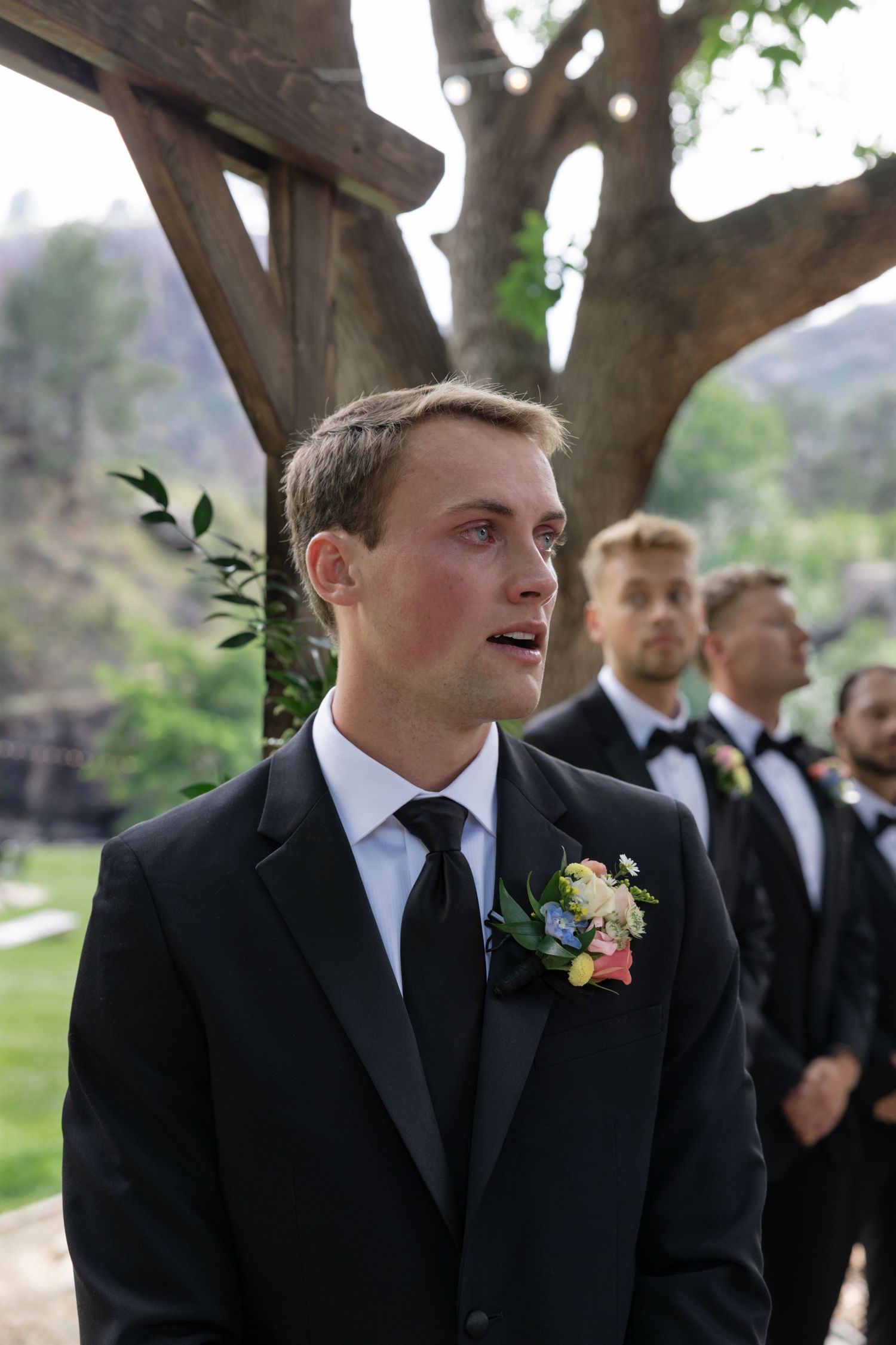 Groom in classic black suit with boutonniere stands during outdoor ceremony.