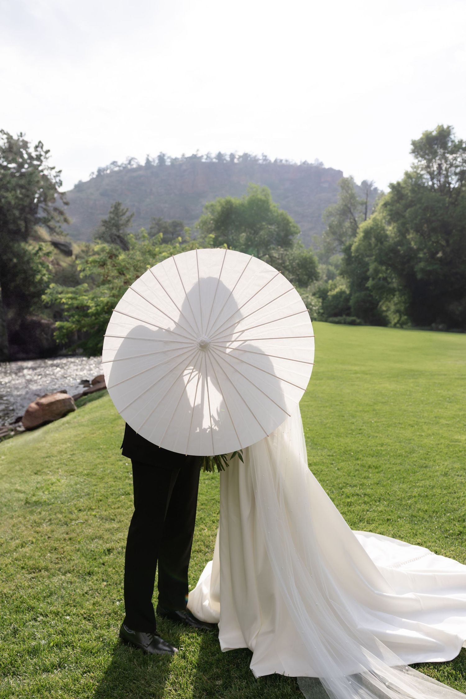 White parasol frames a romantic wedding moment against a mountain backdrop with flowing wedding dress train.