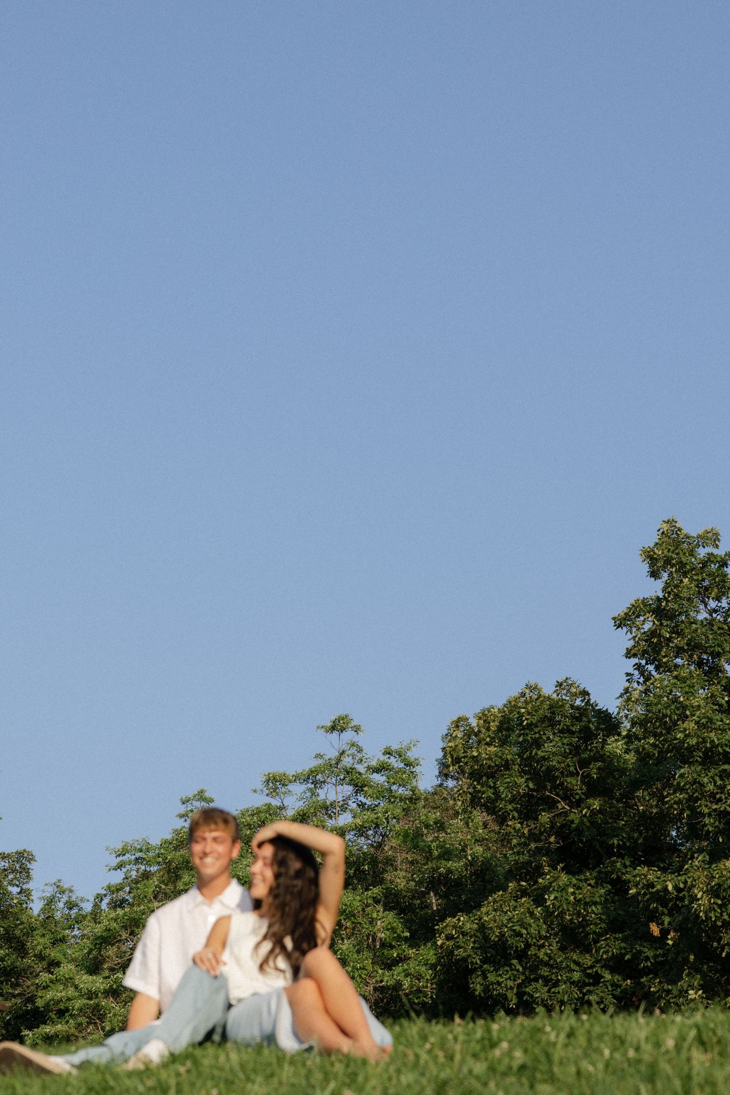 A couple sits on a grassy hillside against a bright blue summer sky and green trees.