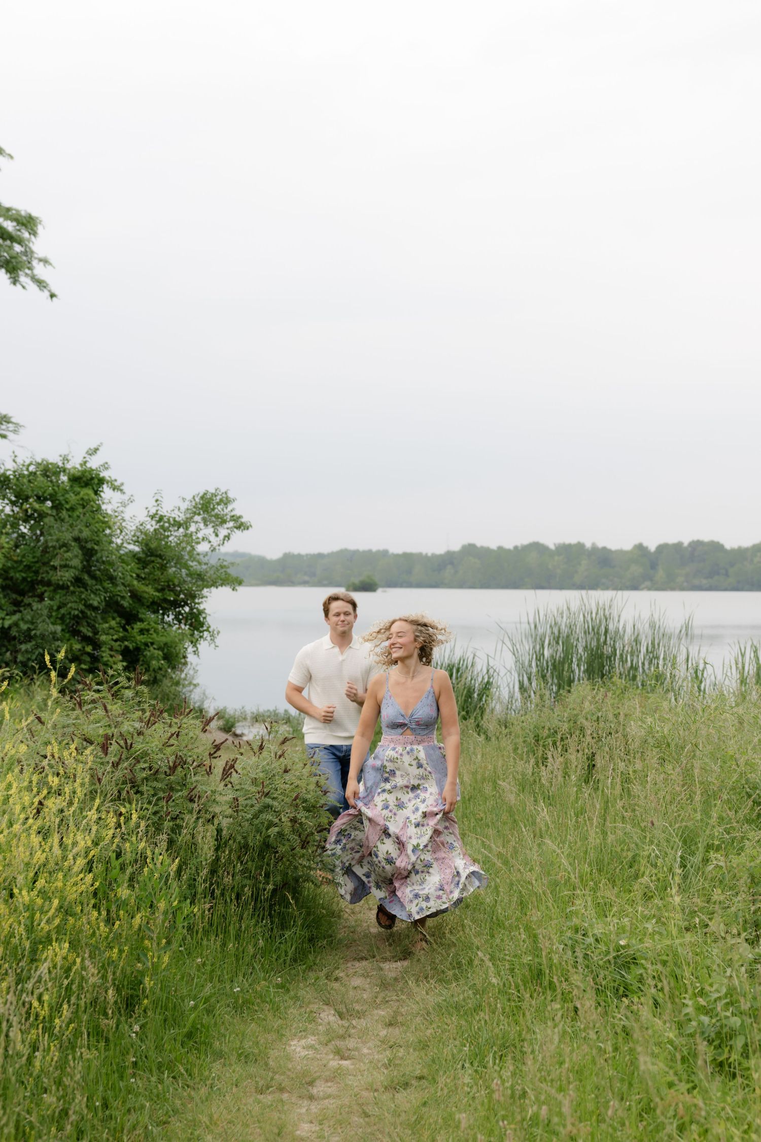 Couple running and playing together through tall grass near a lakeside on a cloudy day.