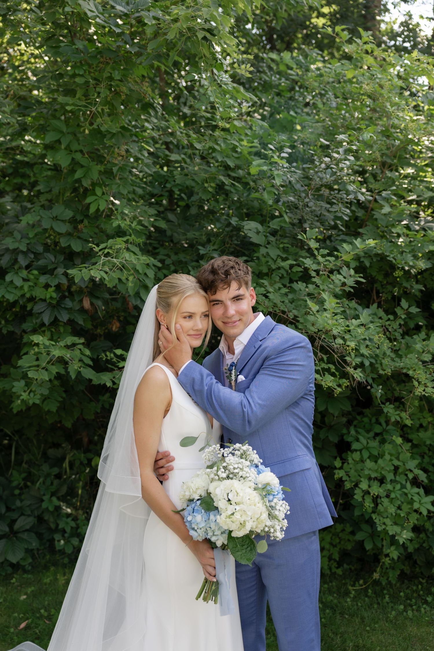 Tender moment between newlyweds with white and blue floral bouquet against green backdrop.