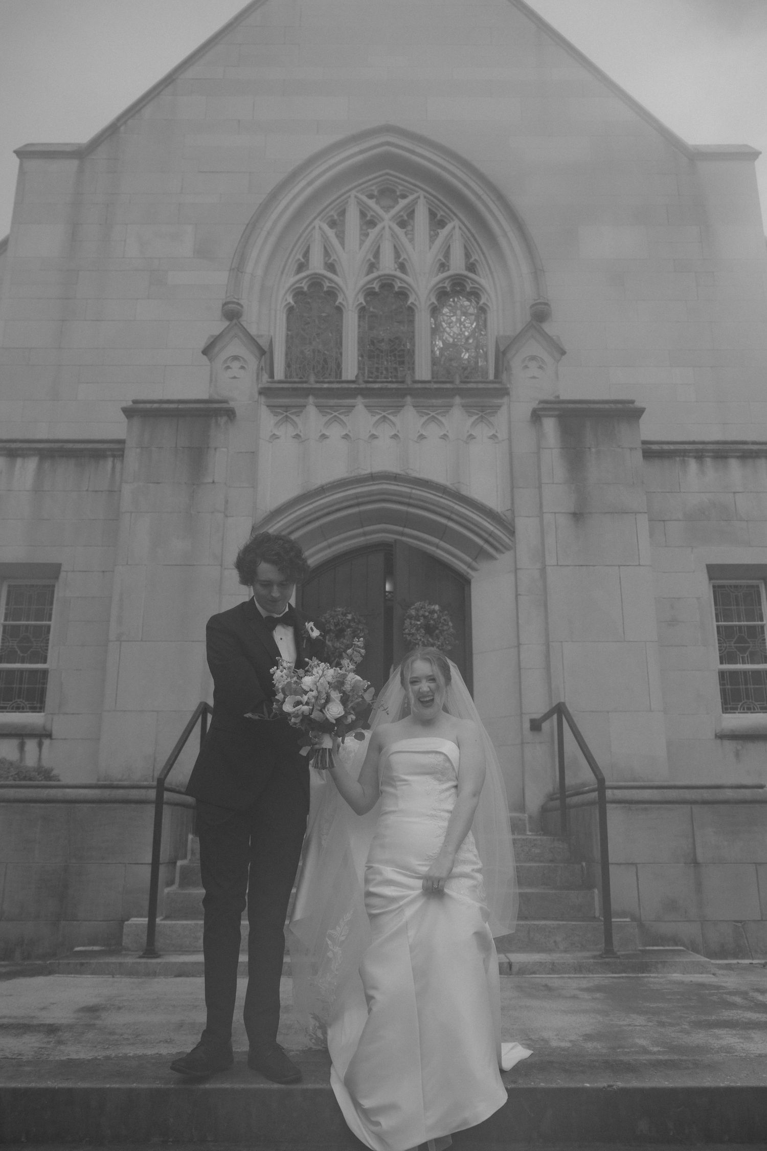 Black and white photo of a couple standing on church steps in formal wedding attire.