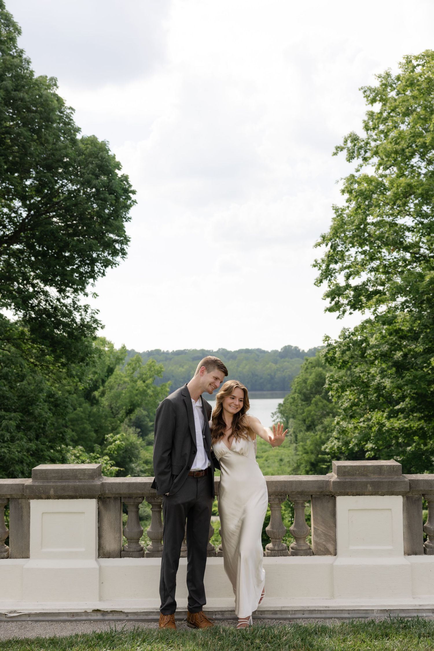 A couple poses against a stone balustrade with a scenic water view in the background.