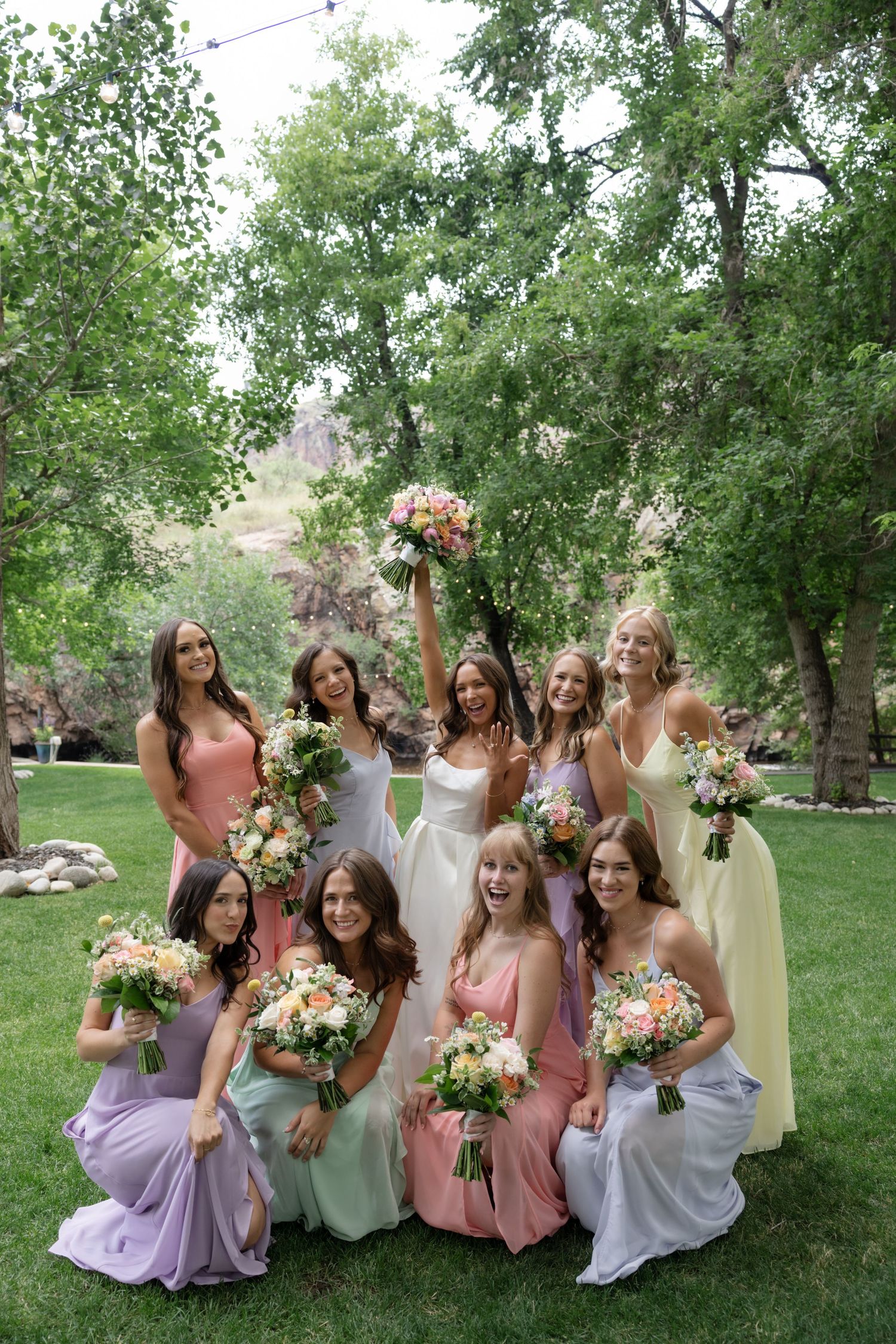 Bridesmaids in pastel dresses posing playfully on grass with bouquets.