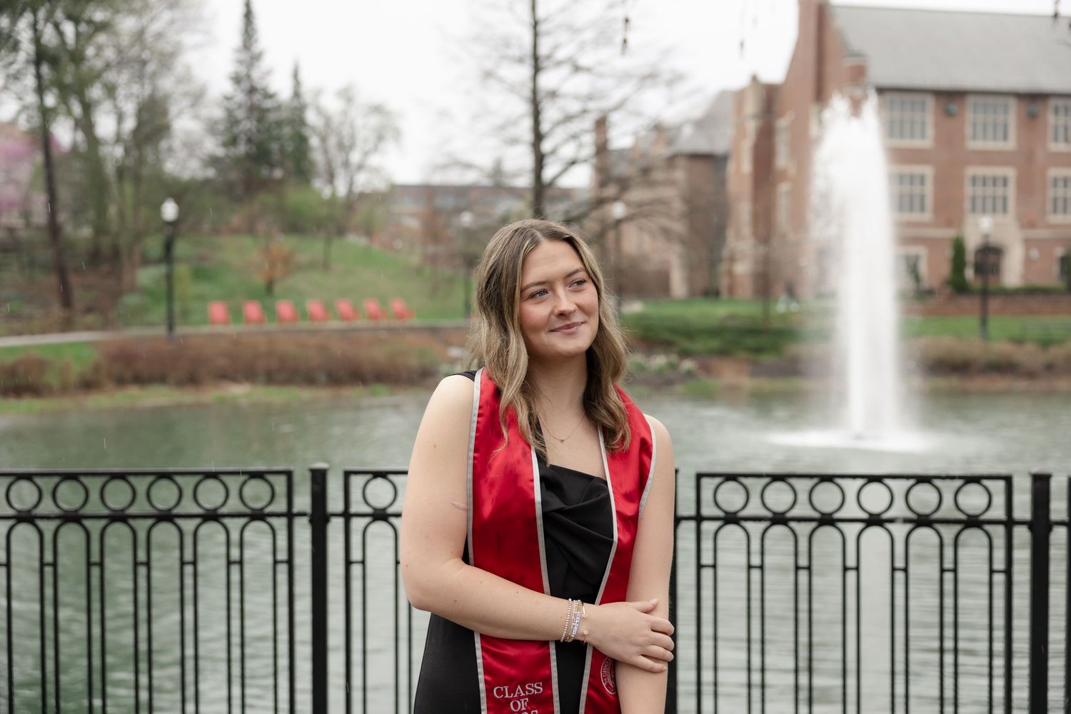 Someone in graduation regalia poses along a wrought iron fence with spring trees in the background.
