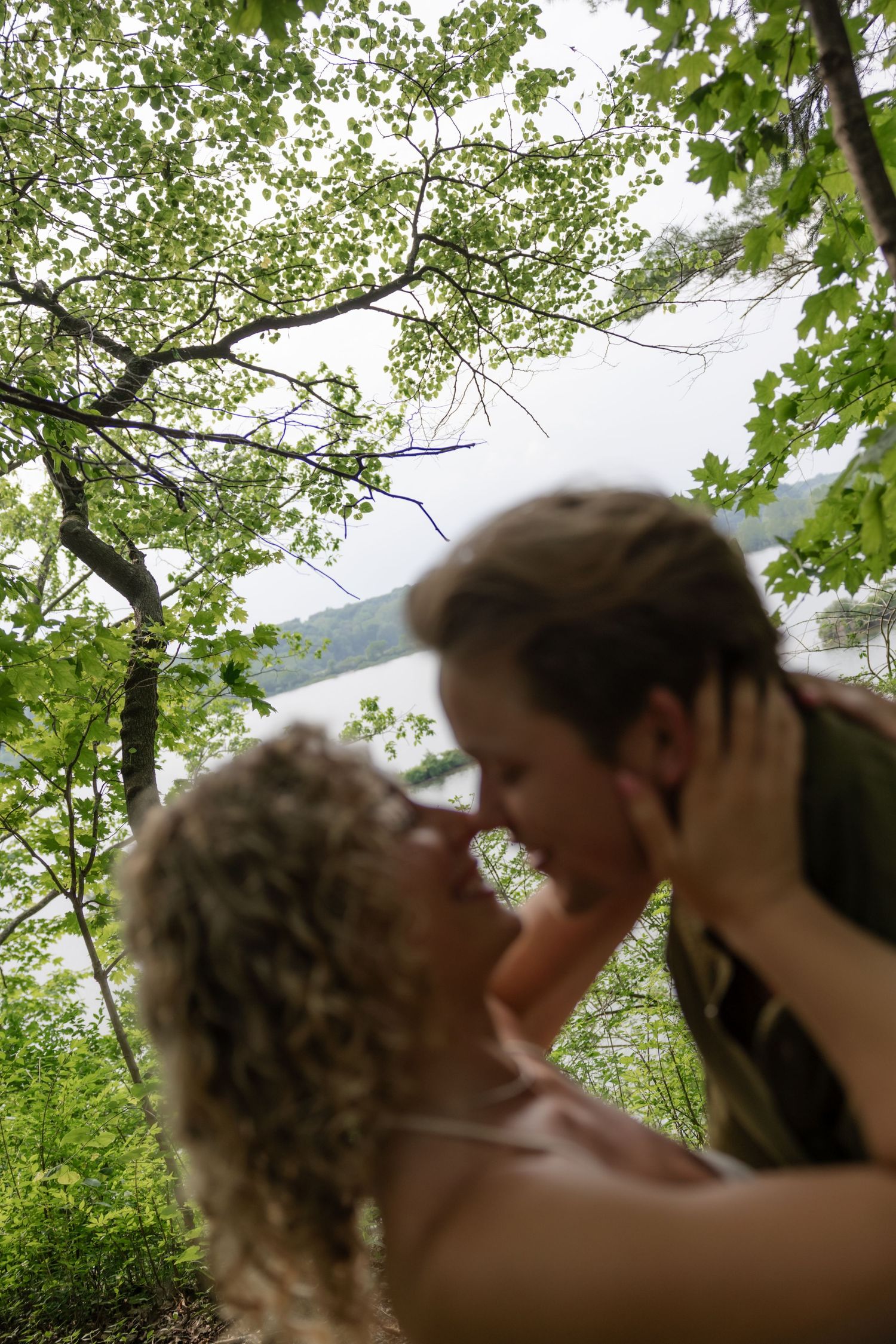 A romantic couple embraces in a natural wooded setting with green foliage in the background.