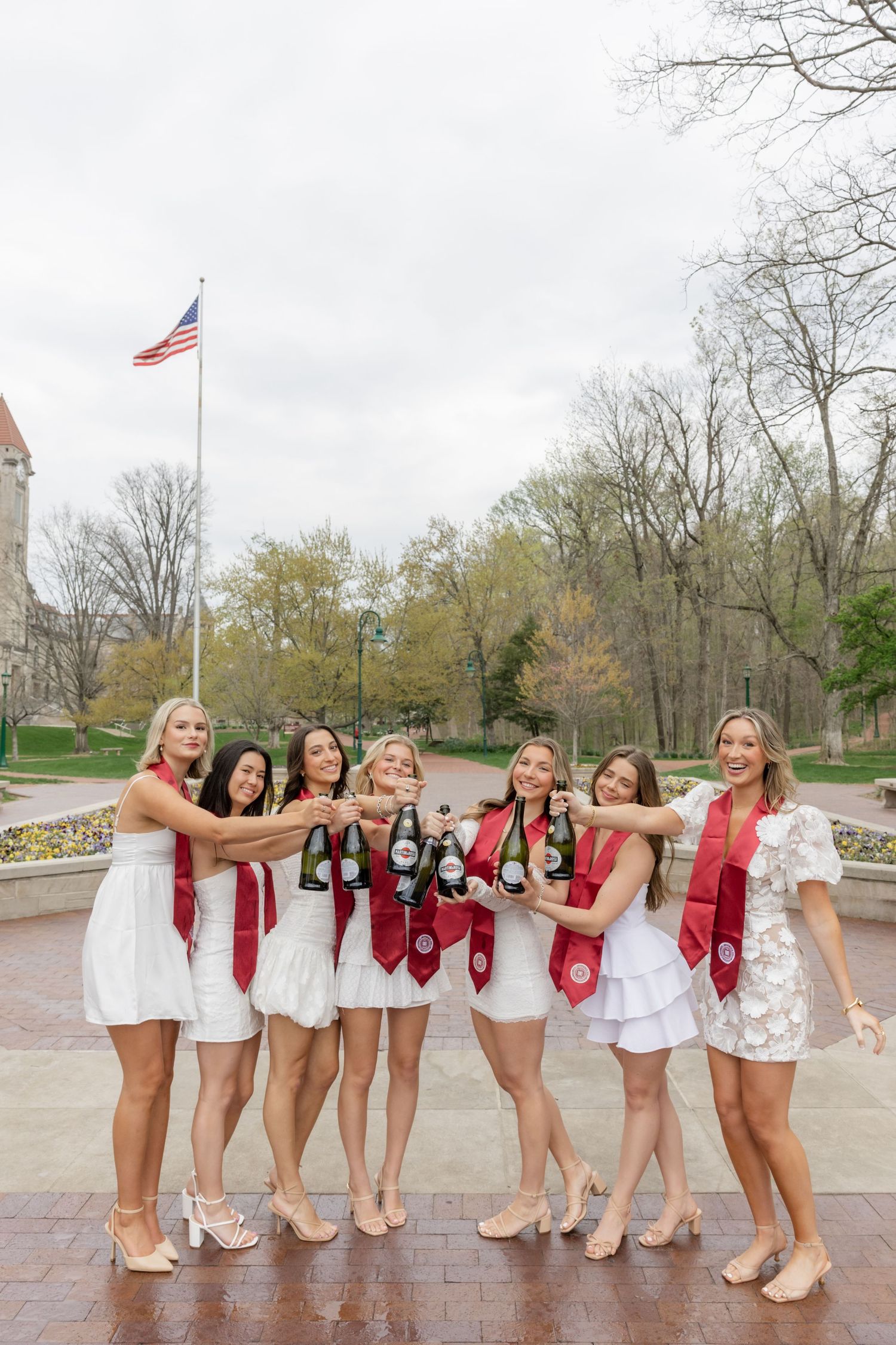 Group wearing white dresses and red sashes pose playfully on brick pavement outdoors.