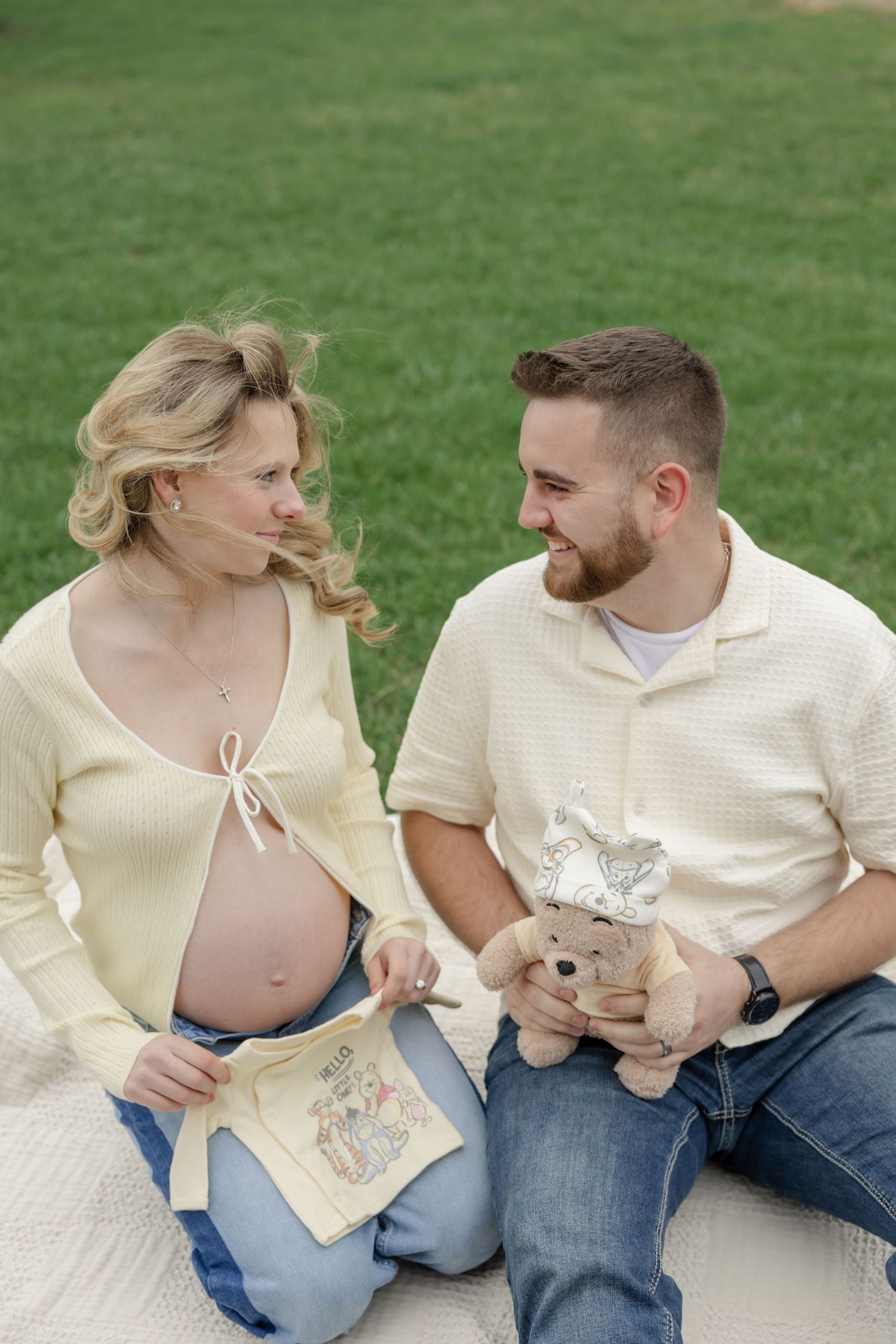 A couple sits on grass looking at each other while wearing casual cream colored outfits outdoors.