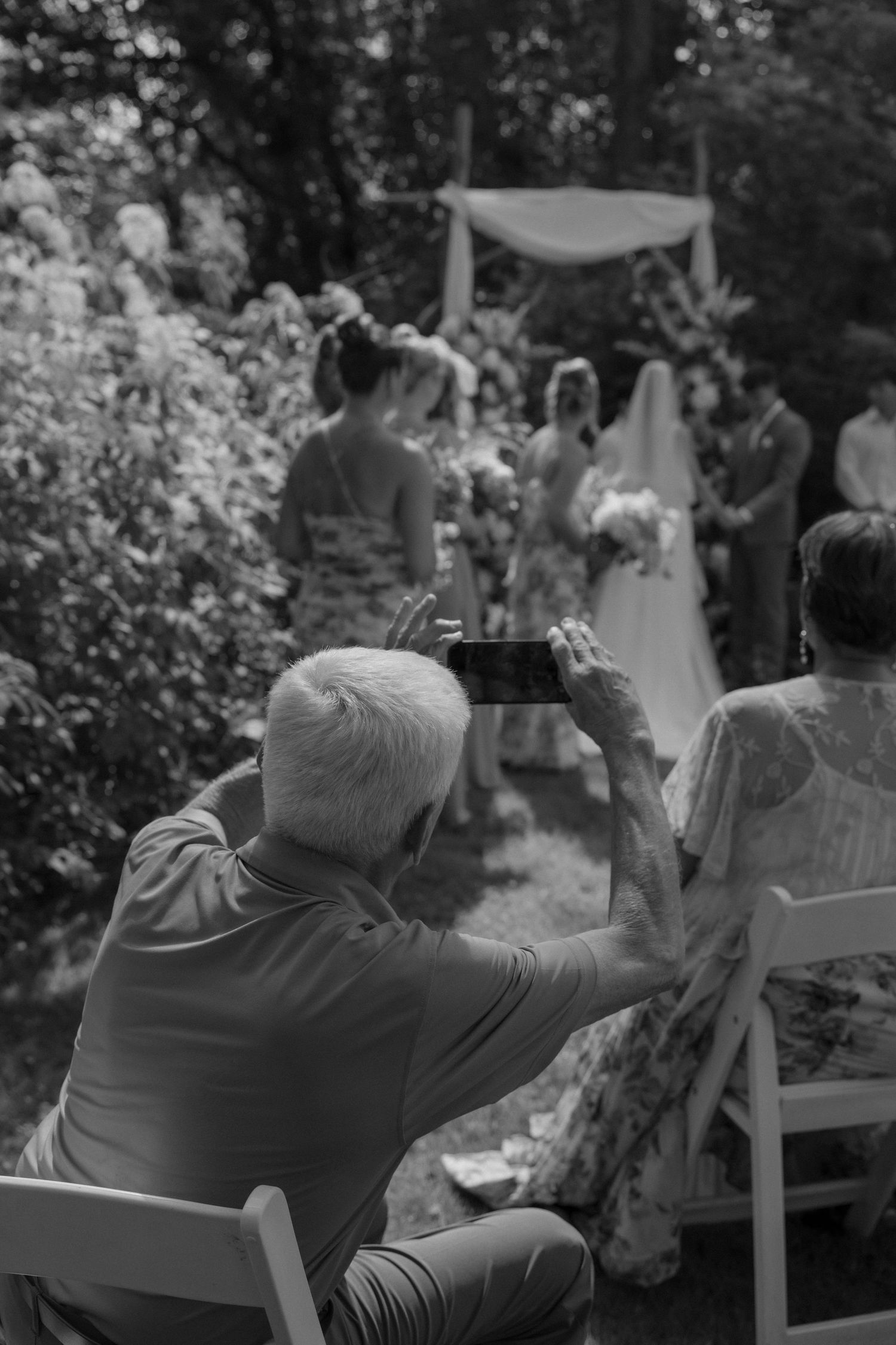 Wedding guests seated in white chairs watching an outdoor ceremony in black and white.