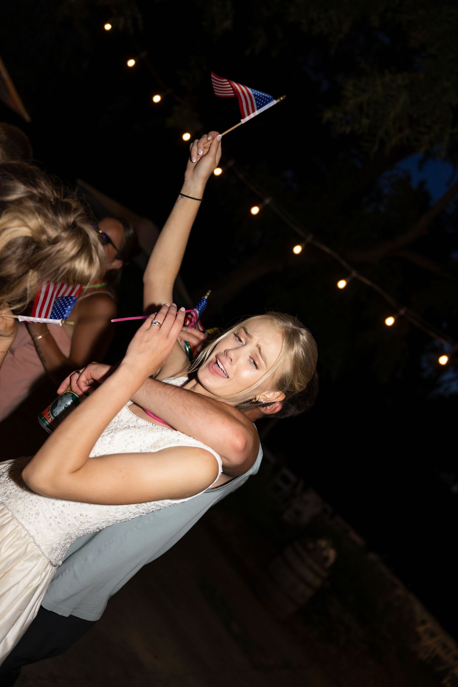 Party guests dancing with small American flags during a nighttime celebration with string lights.