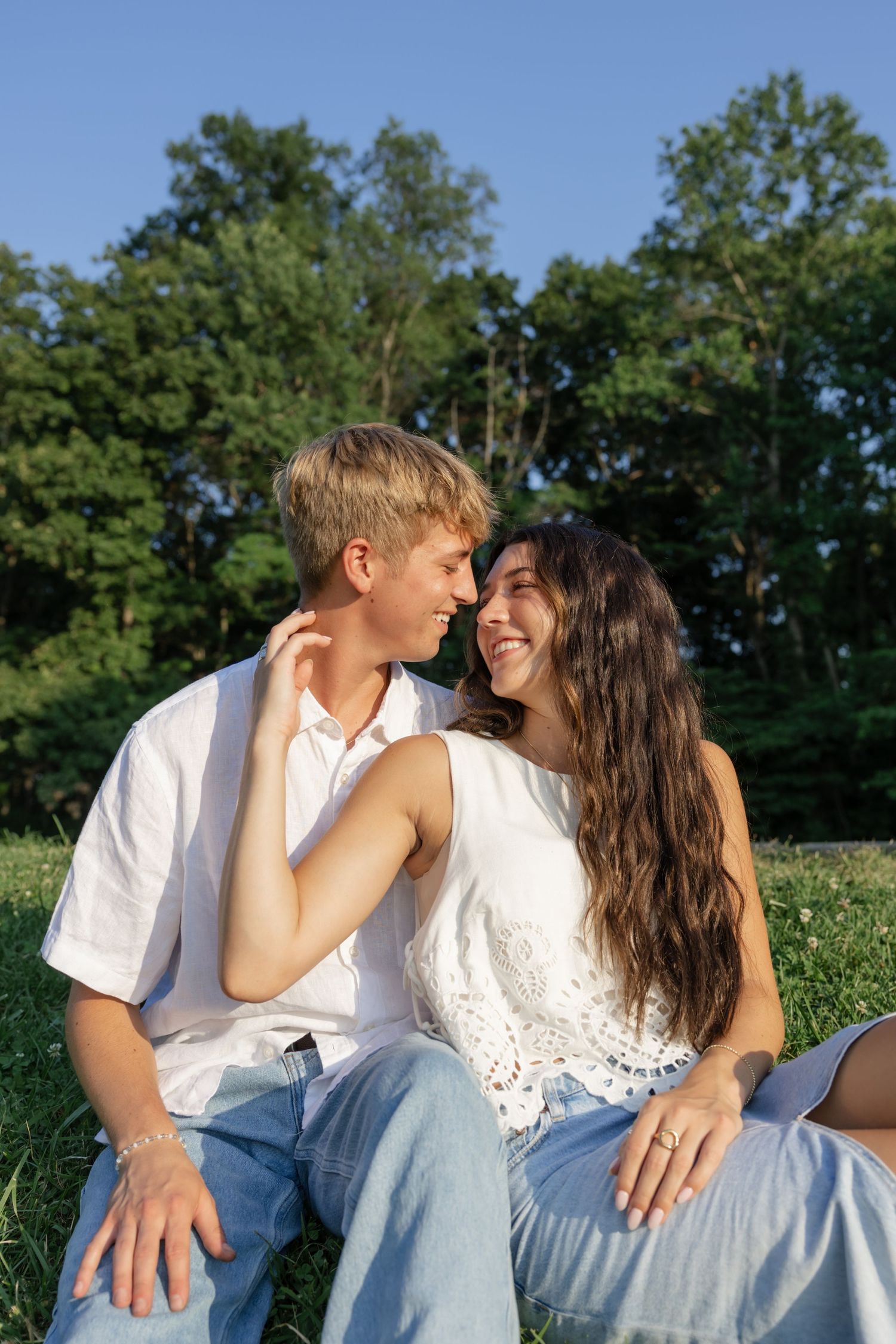 A couple in white clothing shares a tender moment while sitting on grass with trees behind them.