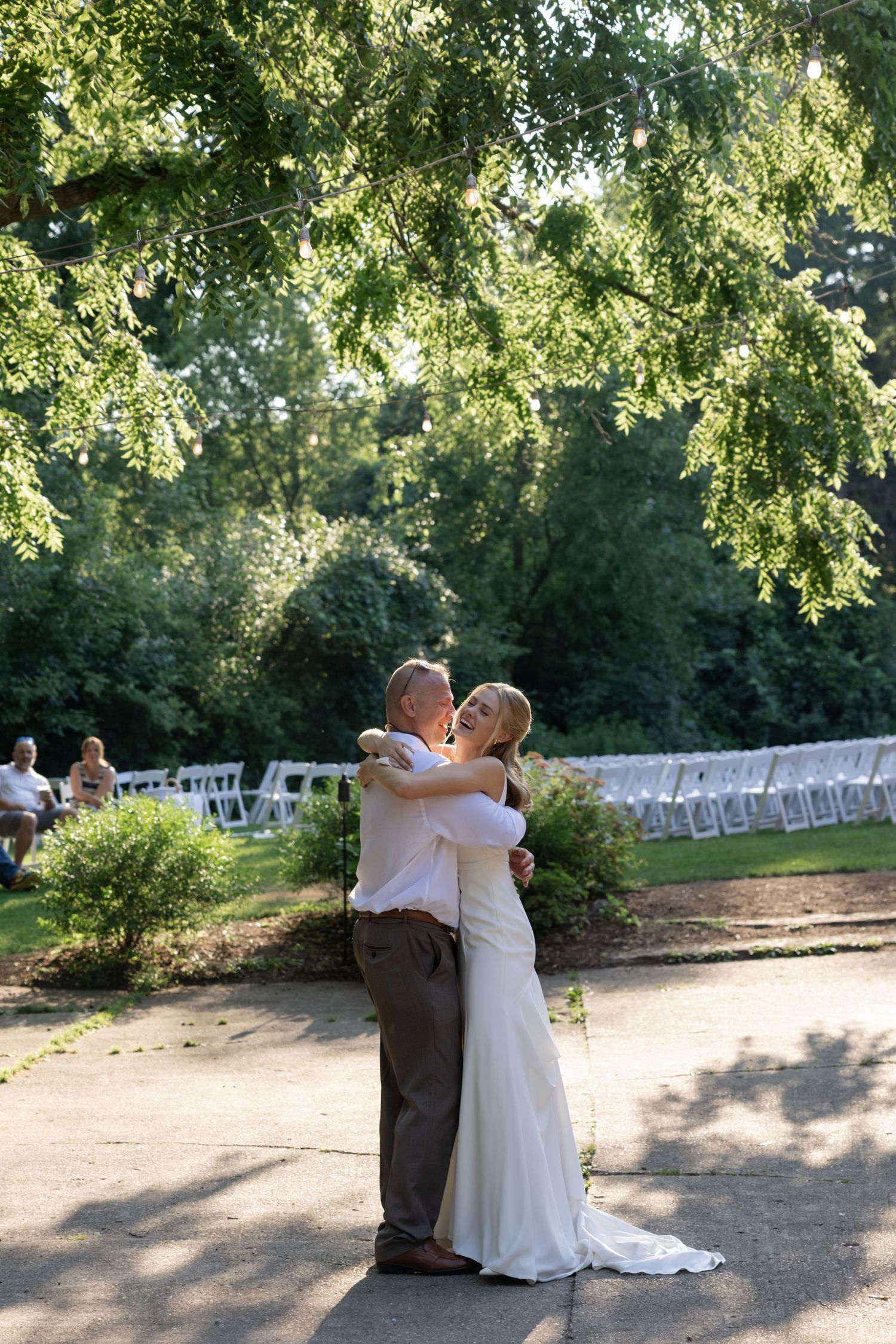 A romantic wedding moment captured outdoors with rows of white chairs and sunlit trees overhead.