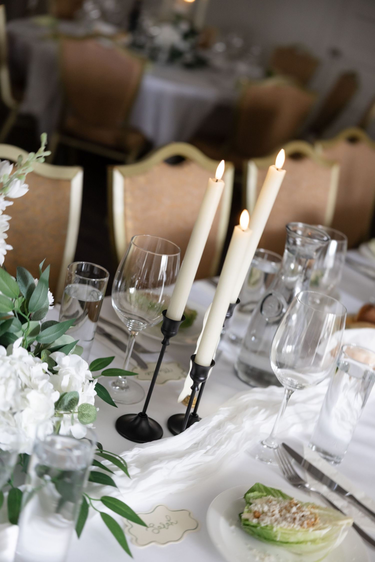 Elegant wedding table settings with white candles, glassware, and green foliage decorations.