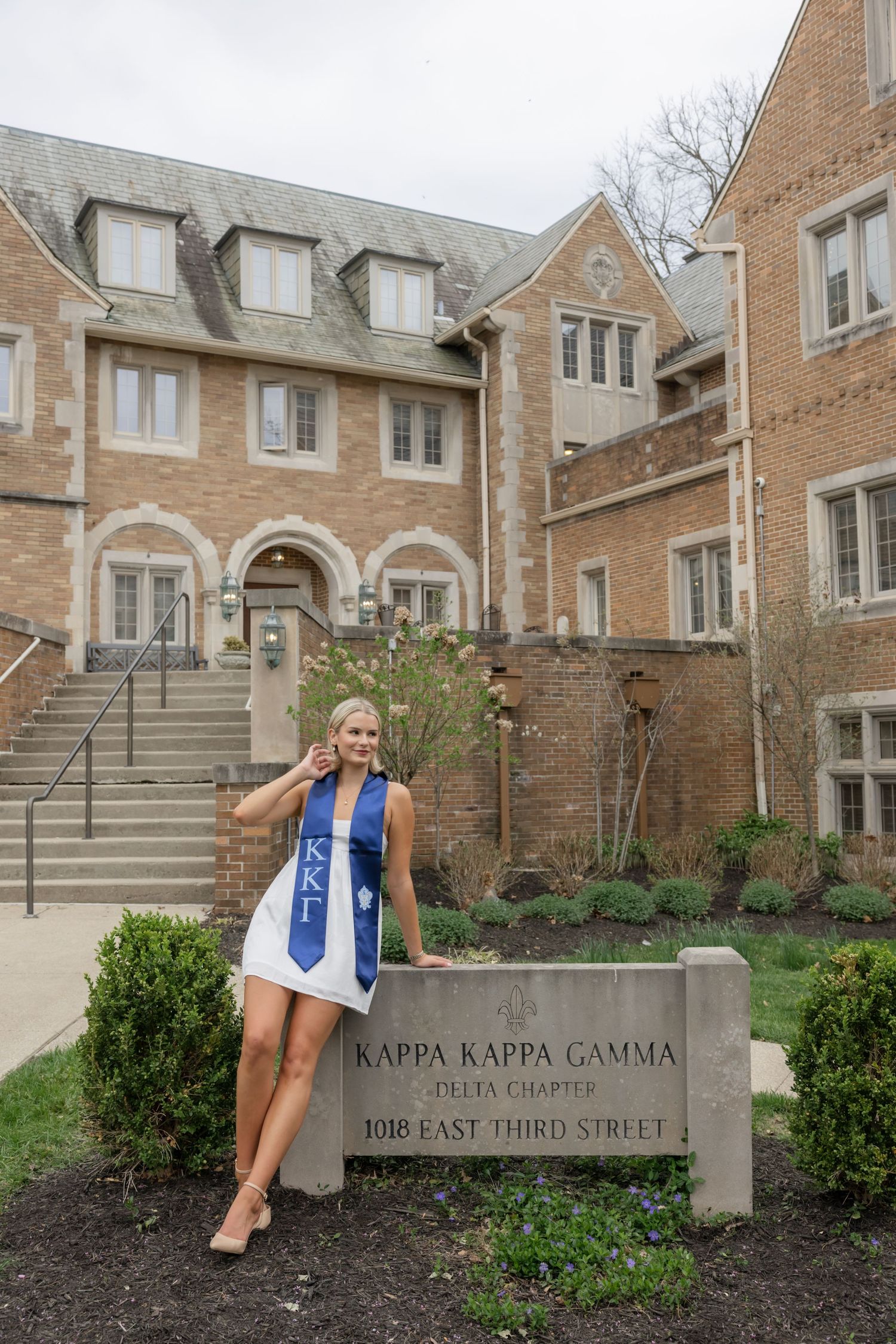 Graduate poses by Kappa Kappa Gamma sorority house sign in white dress with blue sash.
