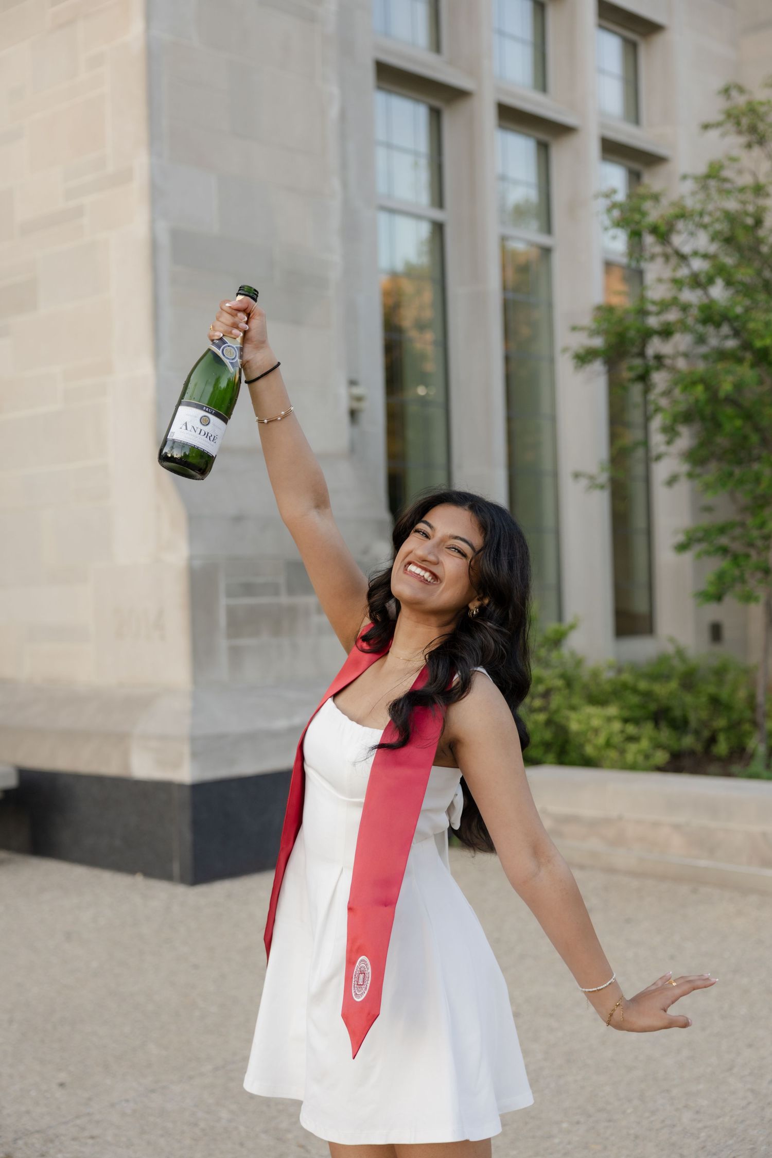 Person in white dress posing with champagne bottle against stone building exterior.