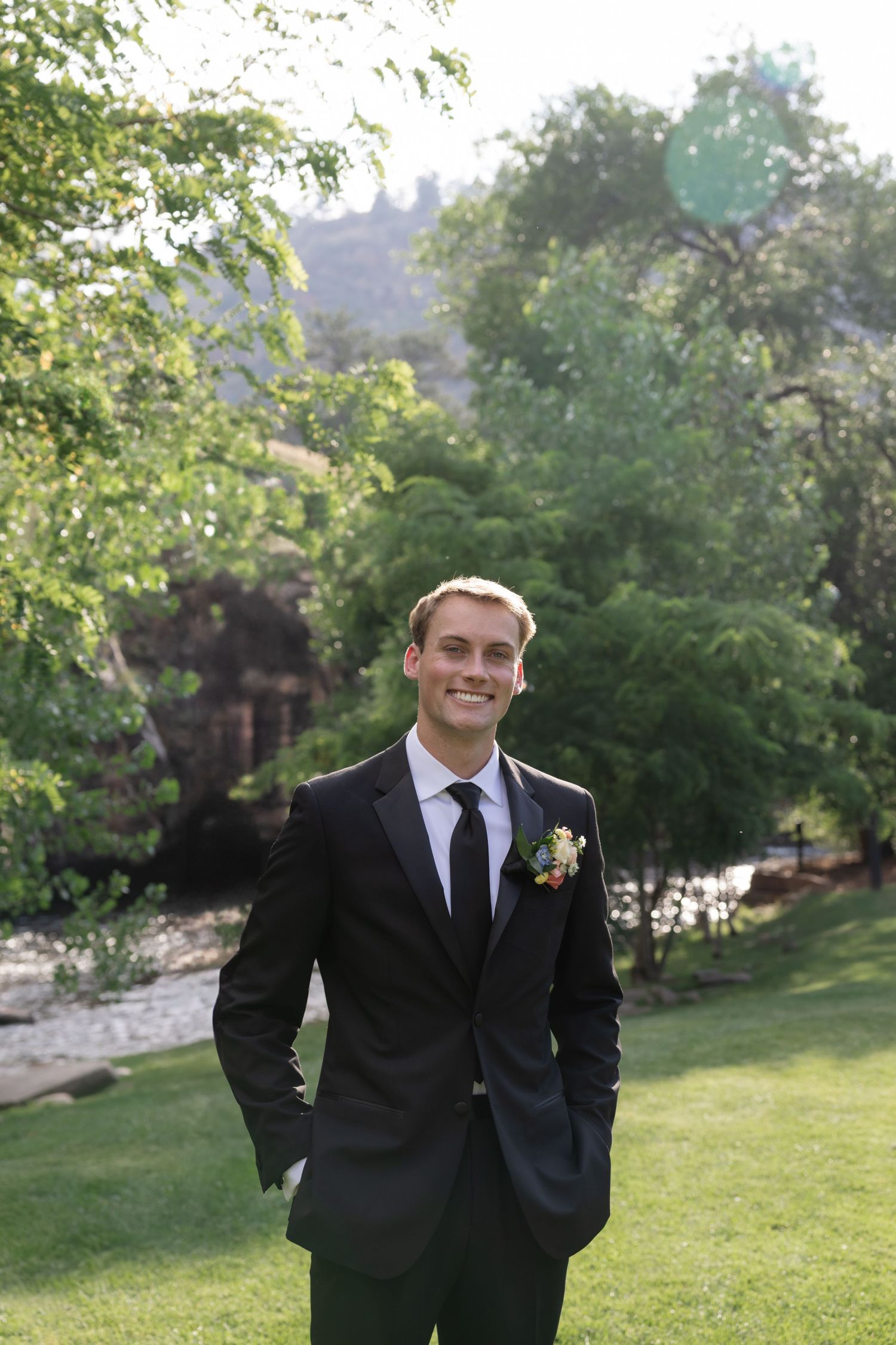 Groom in classic black suit poses in garden setting with natural greenery backdrop.