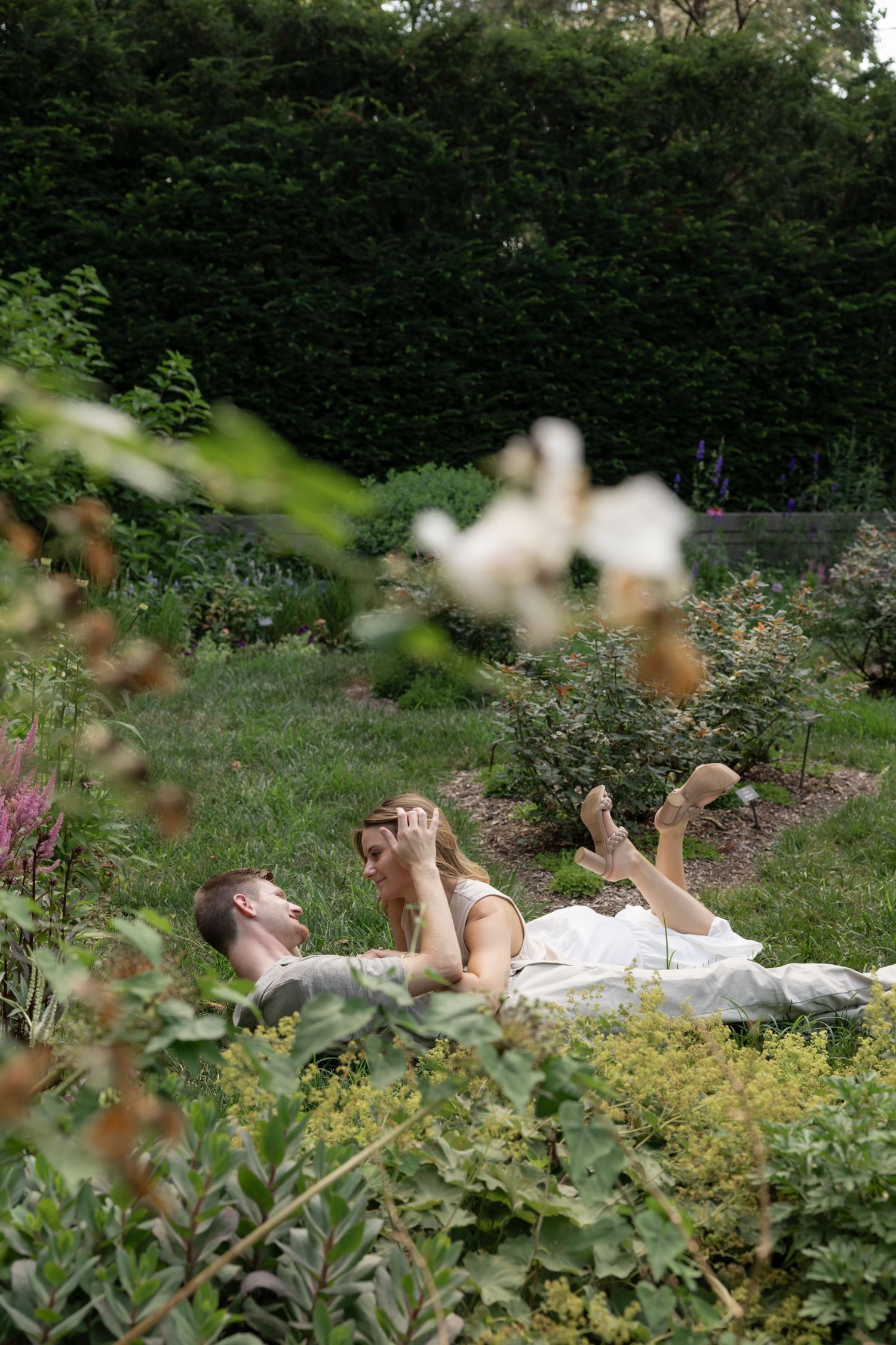 A person lies on a blanket in a garden setting with flowers and hedges.