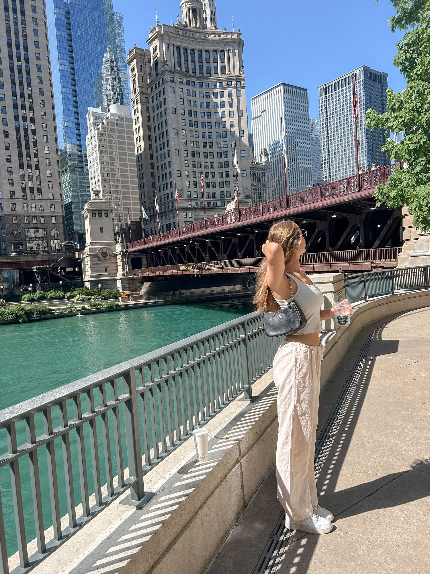 Person in white dress poses along Chicago Riverwalk with skyscrapers and bridge in background.