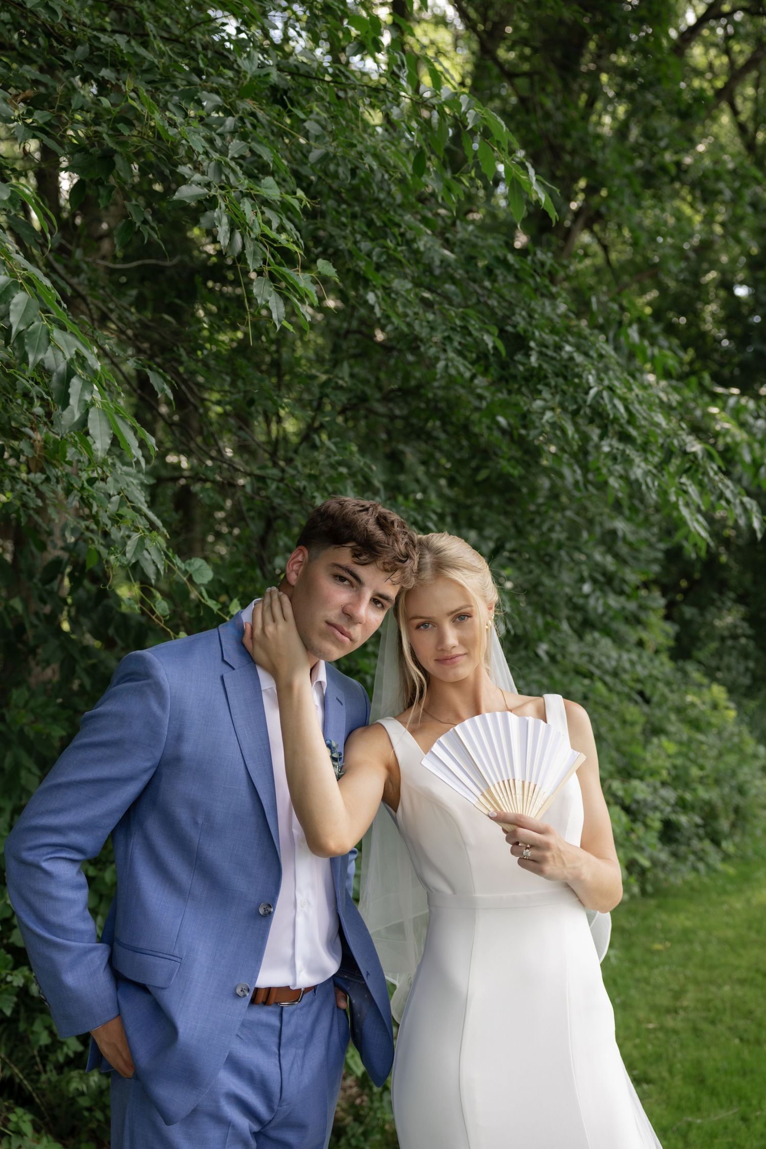 A couple in wedding attire poses on a green lawn with a white folding fan.