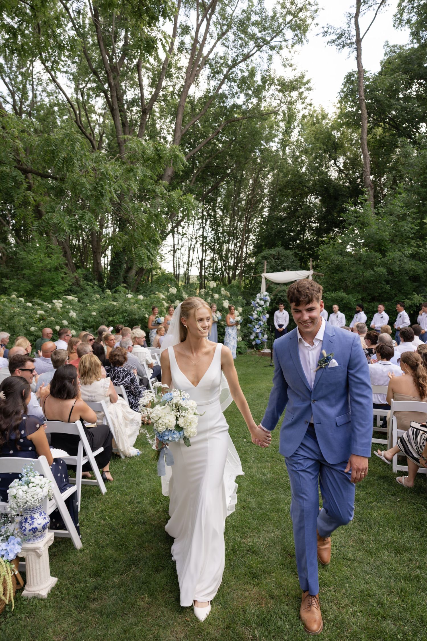 Outdoor wedding ceremony with guests seated on white chairs on a grassy lawn surrounded by trees.