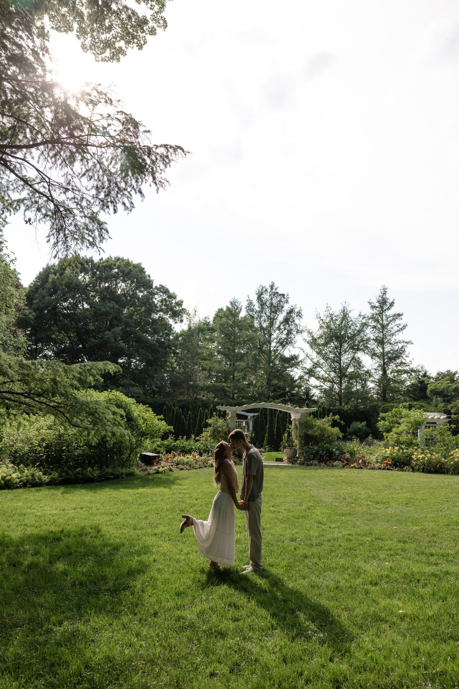 A couple embraces on a sunny lawn with lush gardens and trees in the background.