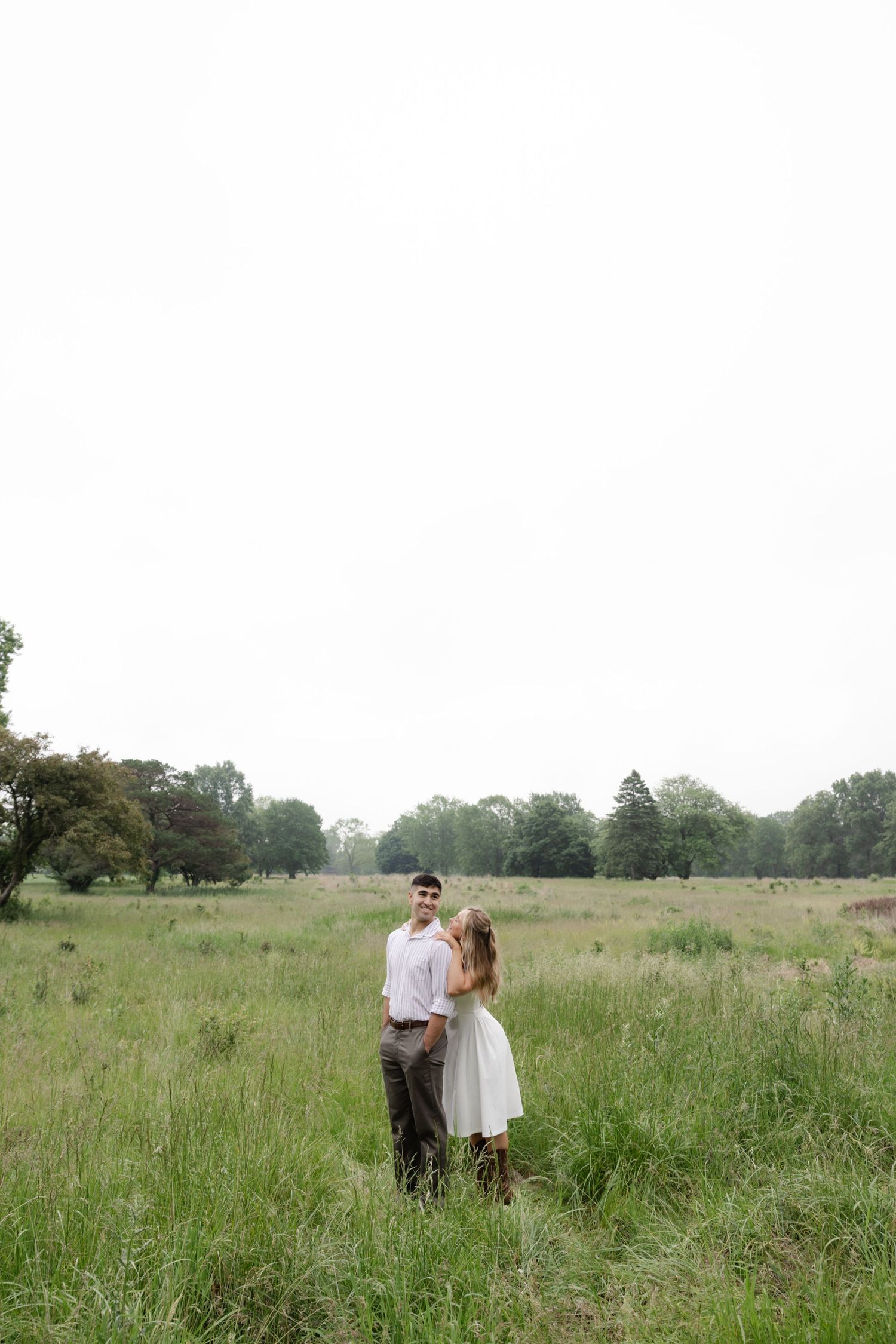 A couple stands together in a vast green meadow surrounded by trees.