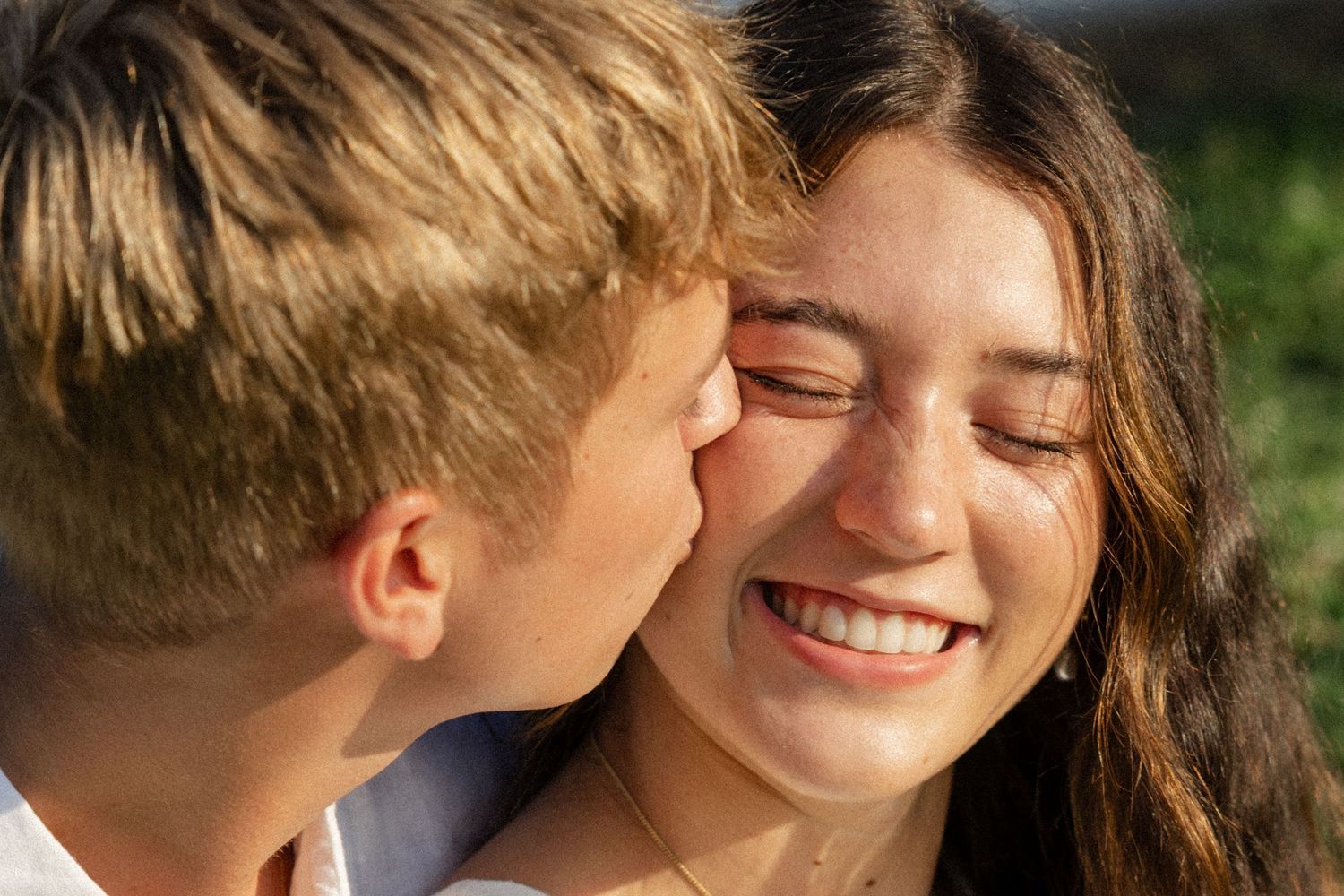 Two people share an intimate moment in warm sunlight with trees in the background.