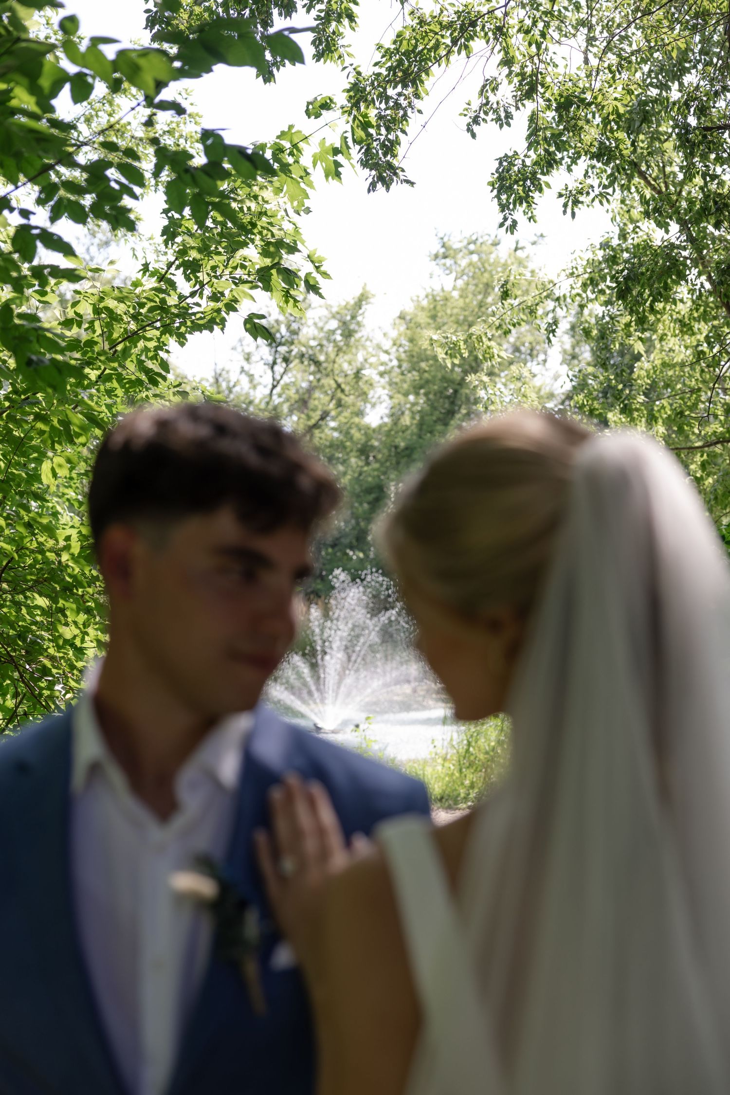 A romantic wedding couple shares an intimate moment under green trees in a garden setting.