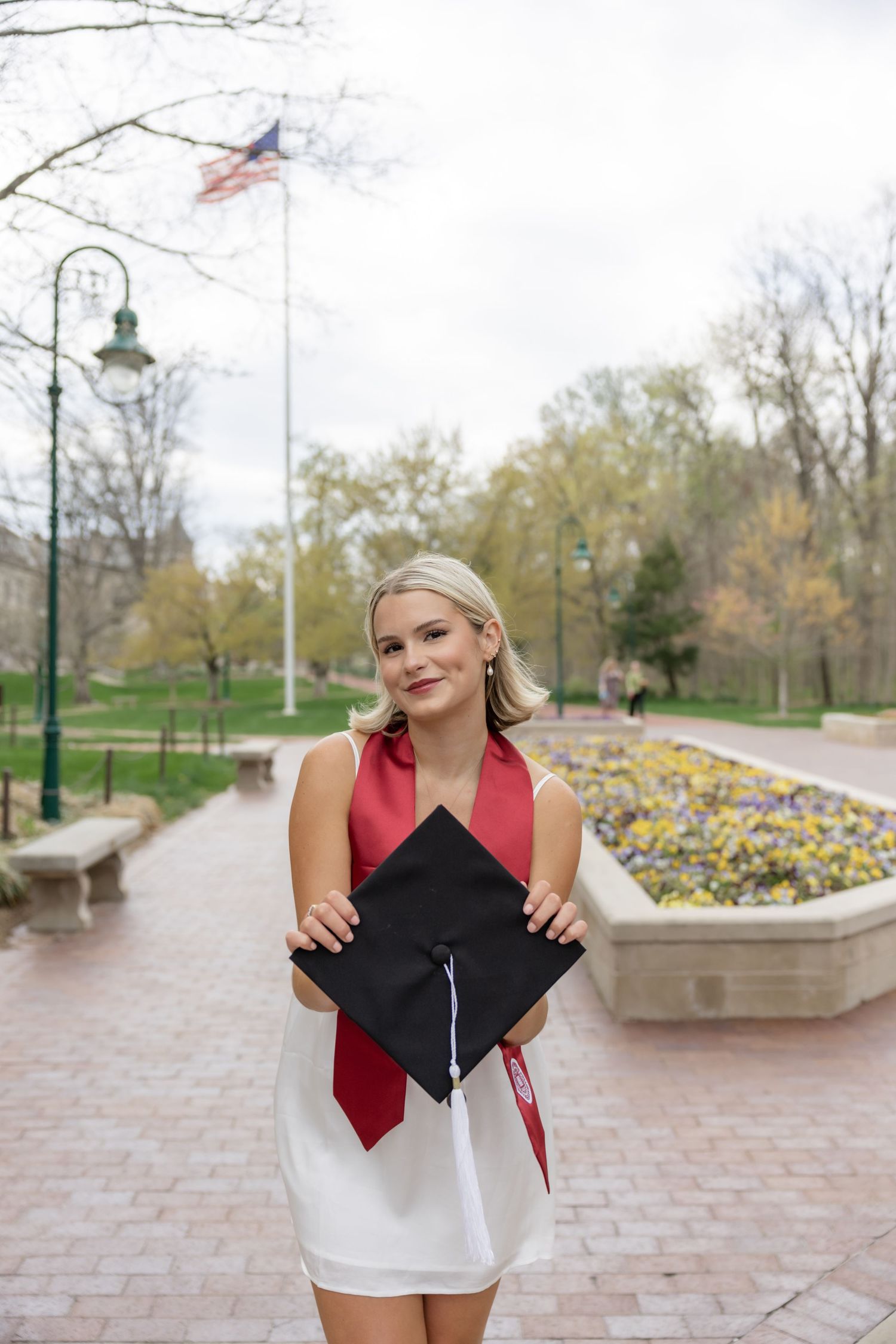 Graduate in white dress and red sash holds graduation cap outdoors with American flag in background.