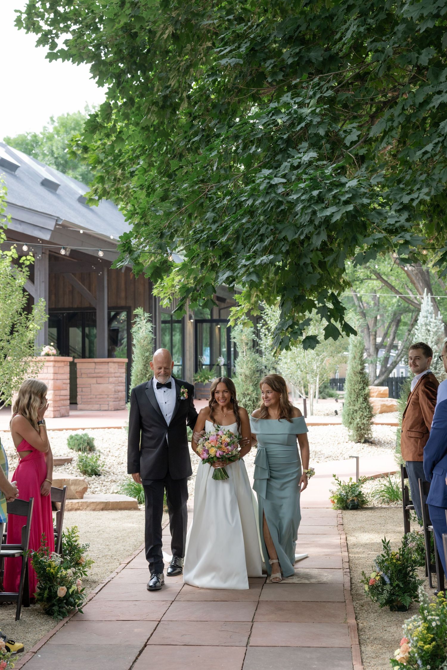Wedding party walks down stone pathway lined with flowers and greenery.