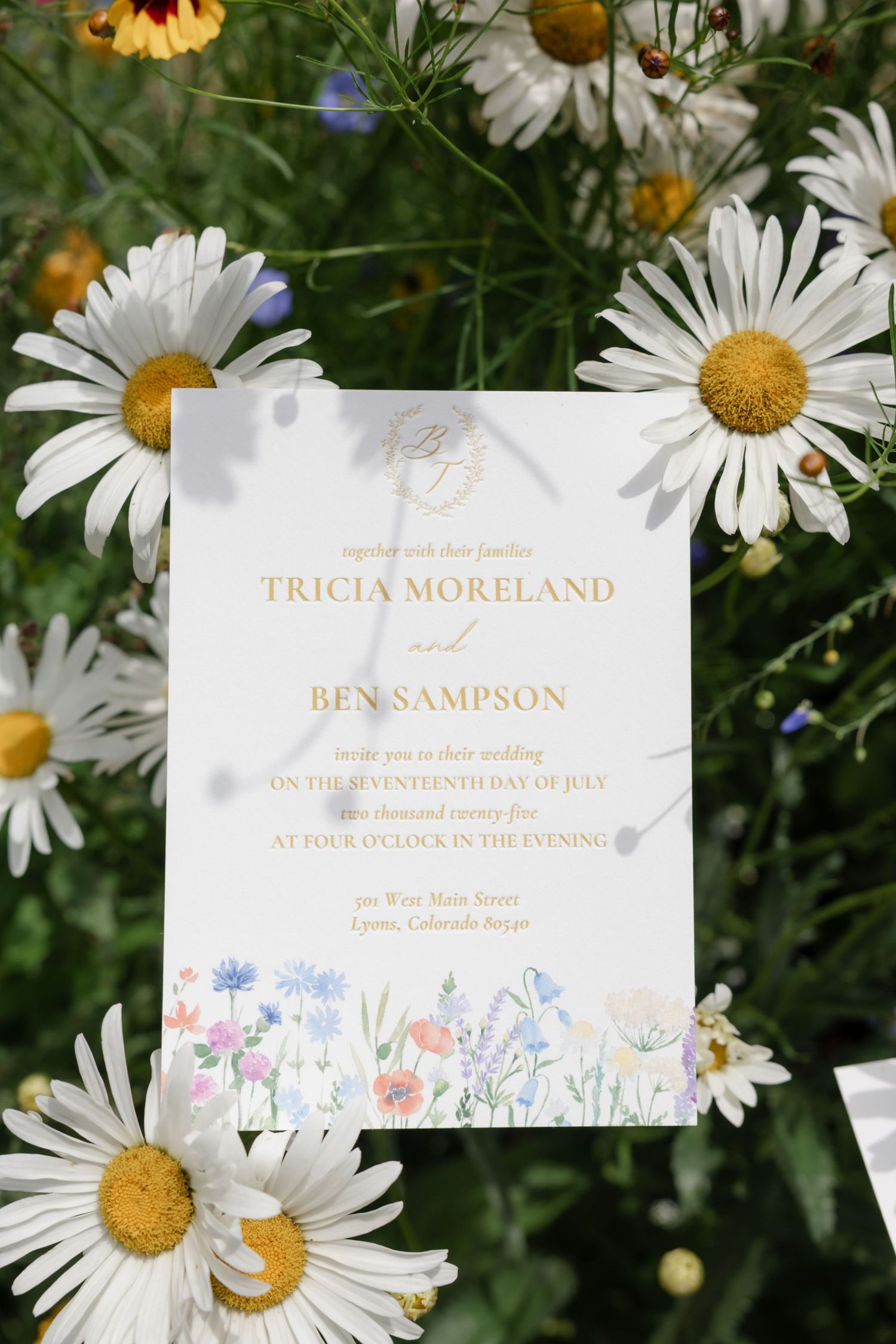 White and gold wedding invitation displayed among blooming daisies and wildflowers.
