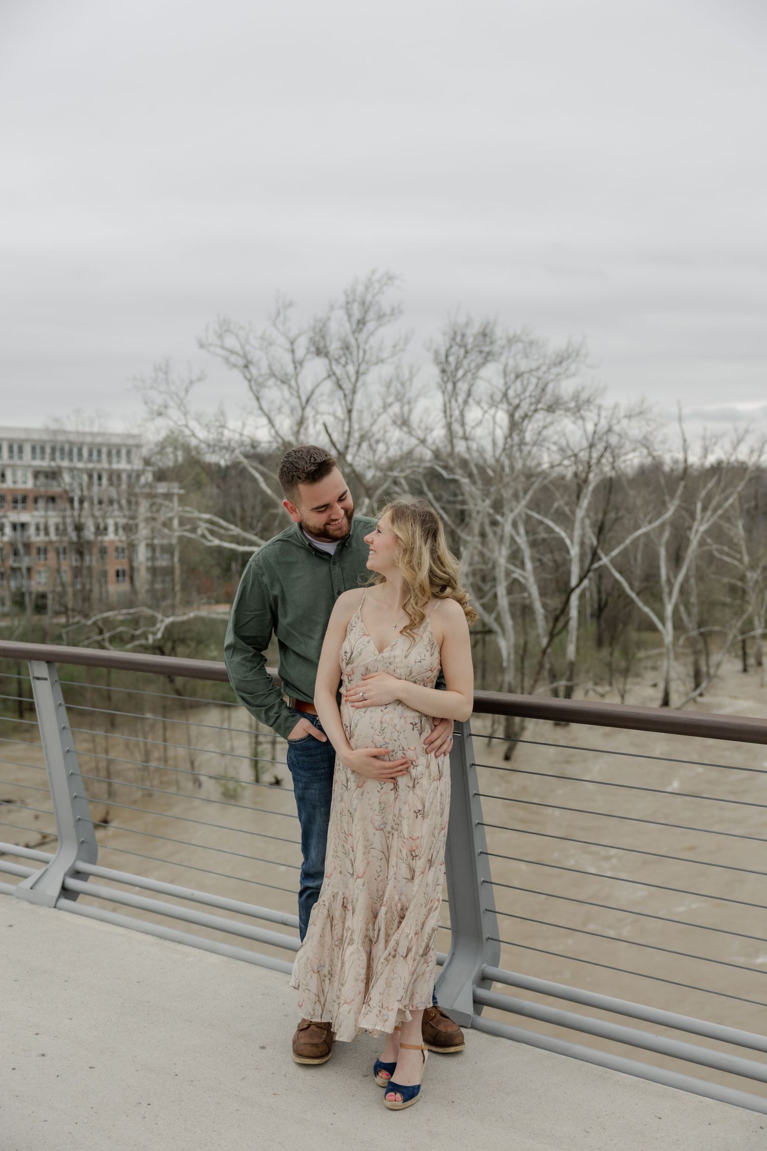 Couple shares an intimate moment on a bridge with apartments and bare trees in background.