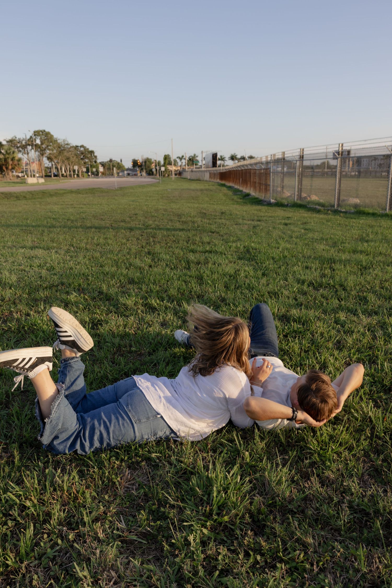 People lying in green grass near airport fence watching planes overhead.