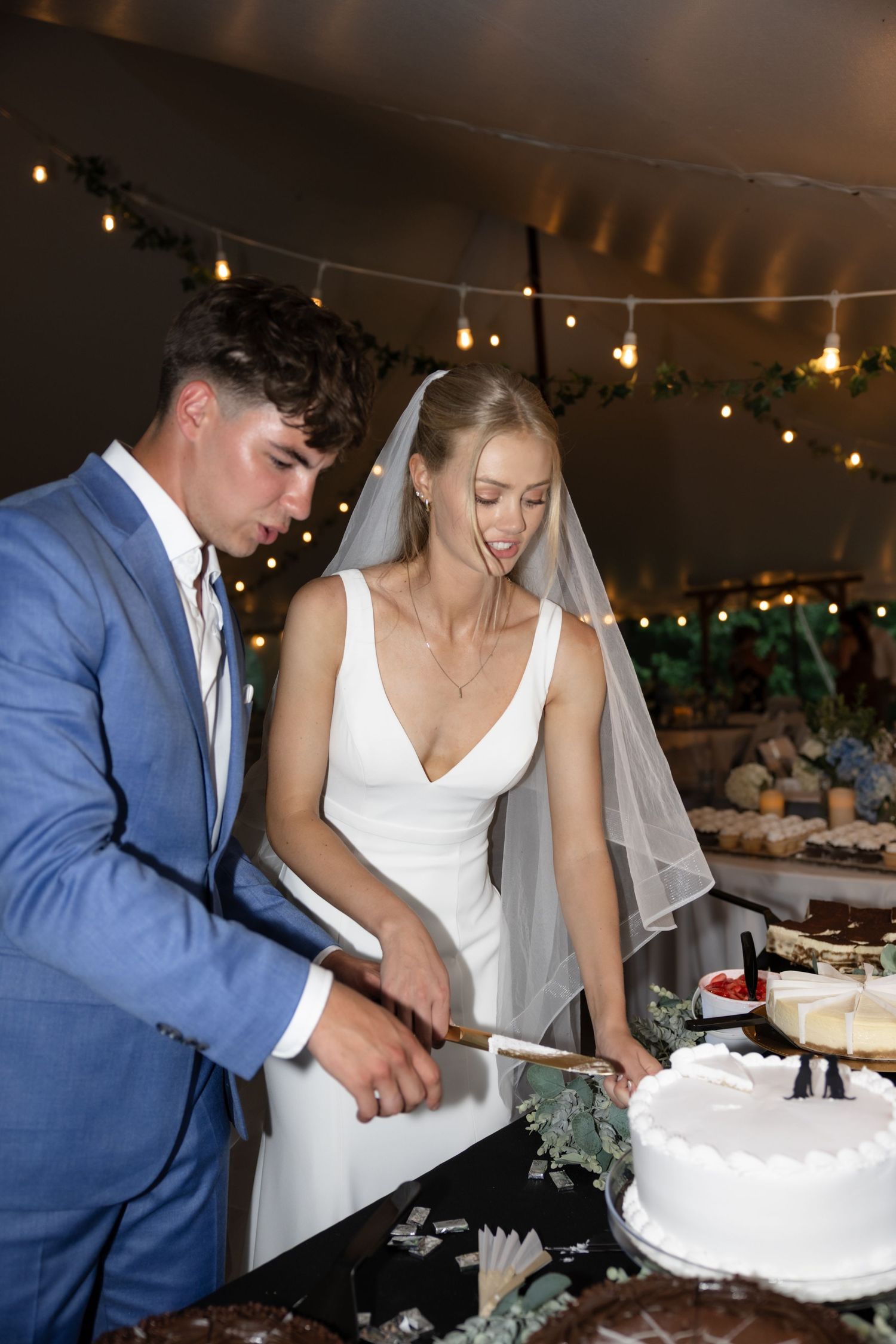 Couple cutting their white wedding cake together under string lights at reception.