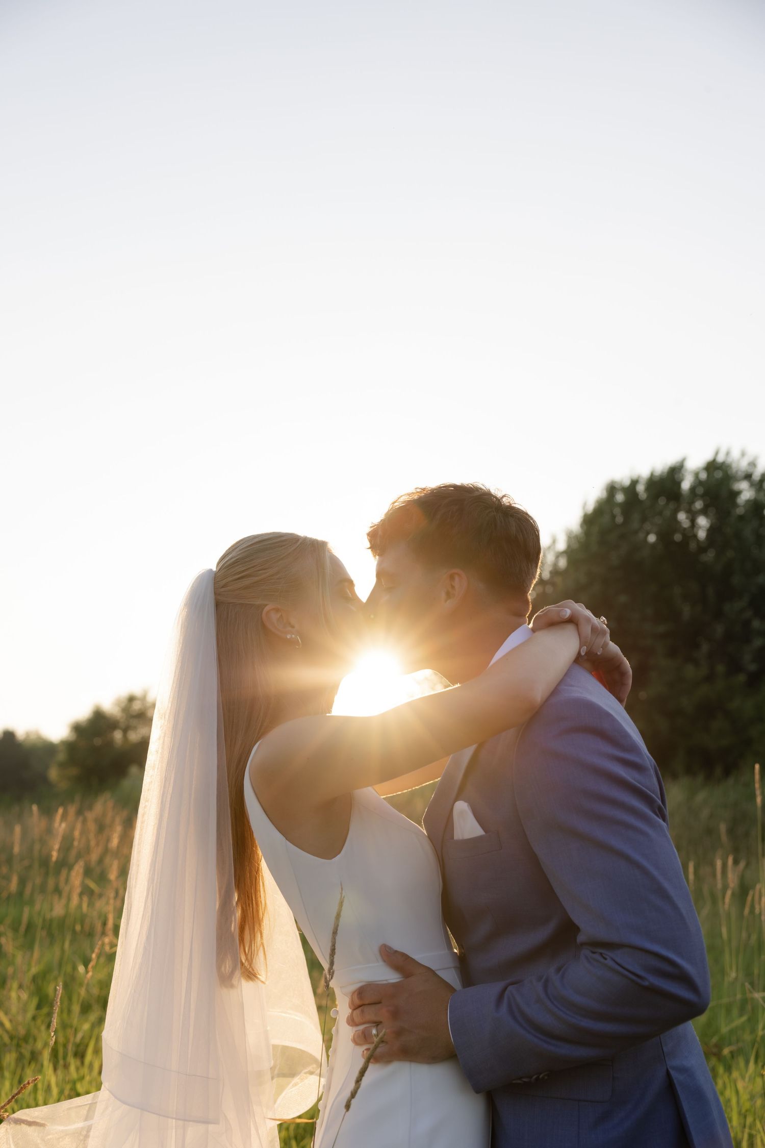 Newlyweds share a romantic sunset kiss in a field with golden light behind them.