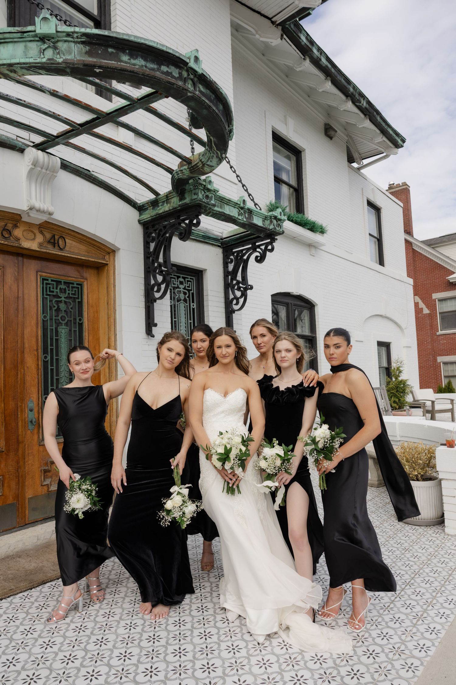 Wedding party in black dresses poses with bride in white gown under an ornate architectural awning.
