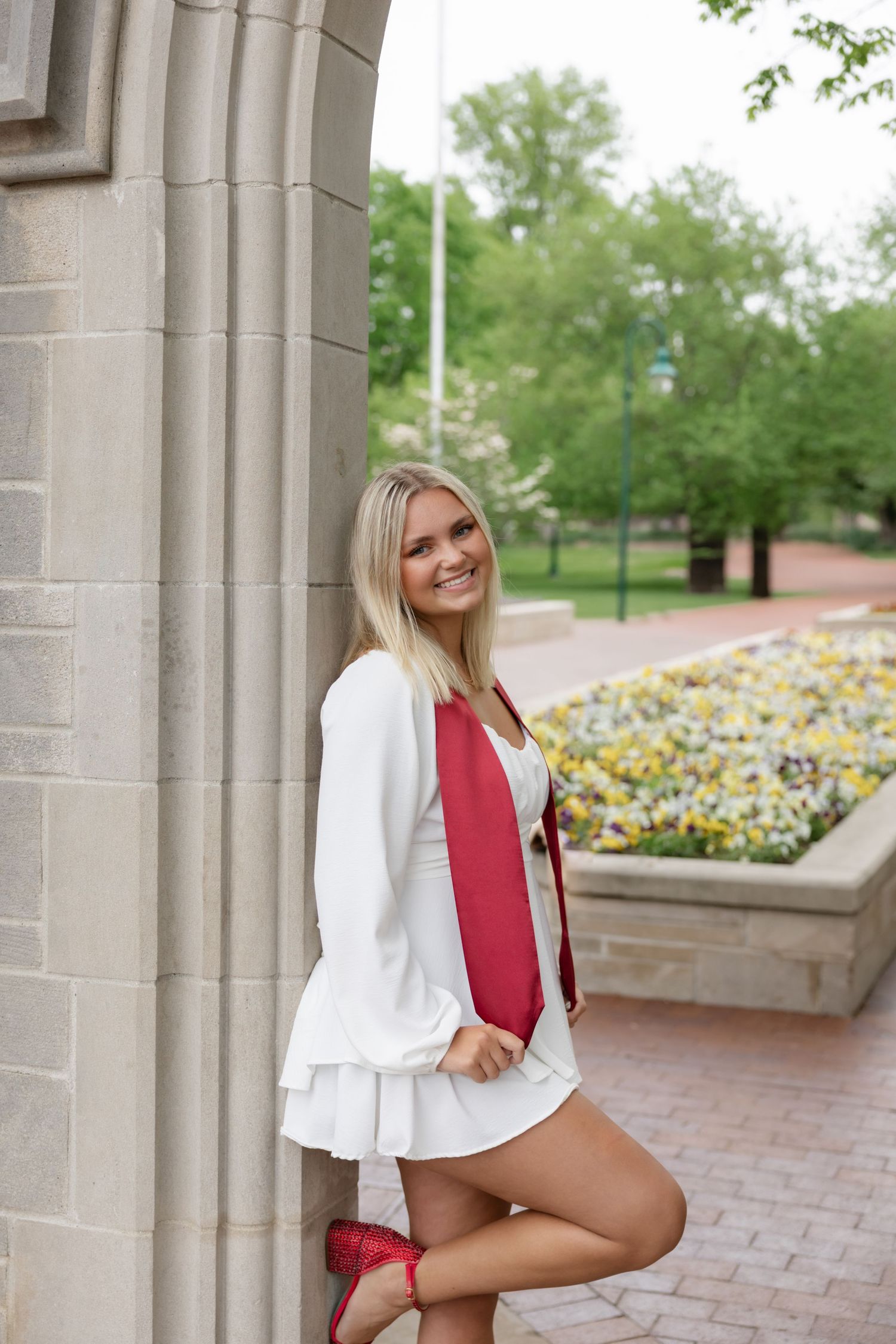 Graduate in white dress and red sash poses by stone column with flower garden in background.